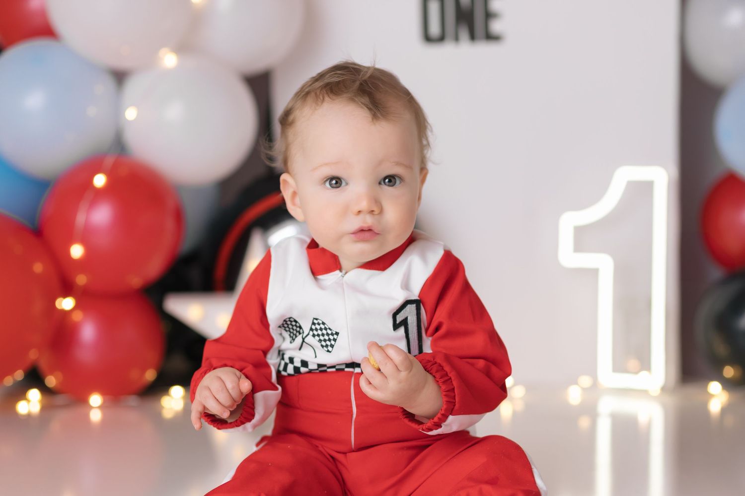 Baby in red outfit sits among festive white and red balloon decorations with glowing number one display.