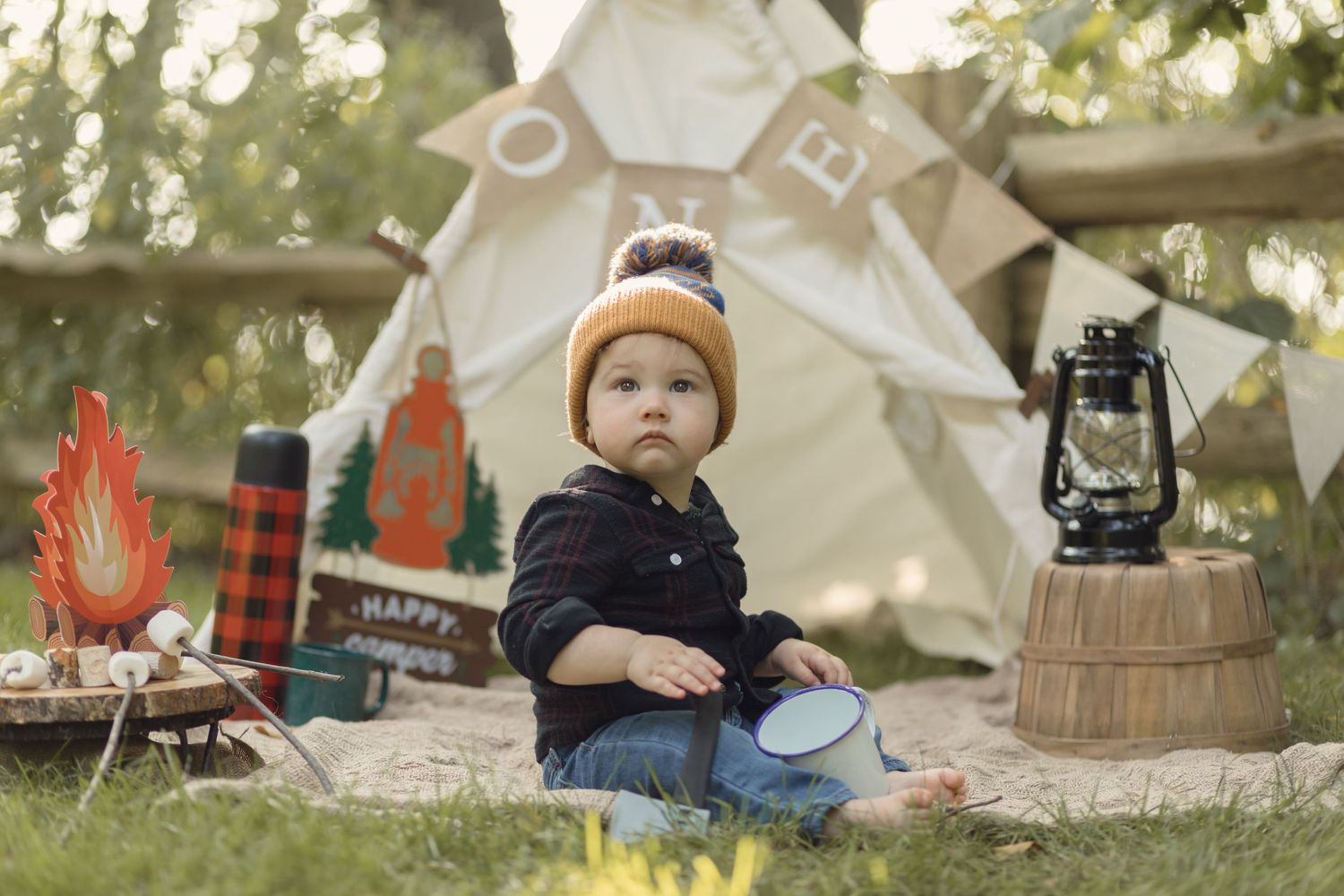 A small child in a winter hat sits near a teepee tent with camping decor including a lantern and faux campfire.