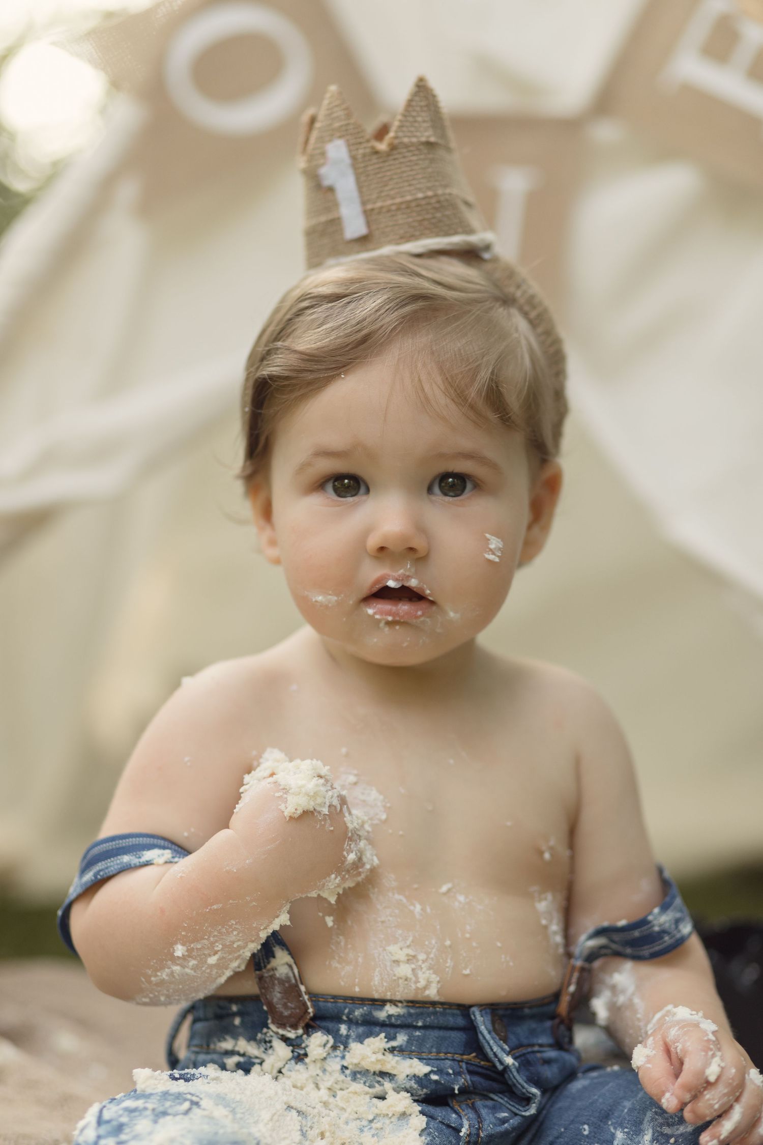 Small child wearing a paper crown and jeans gets messy while playing with cake during a first birthday celebration.