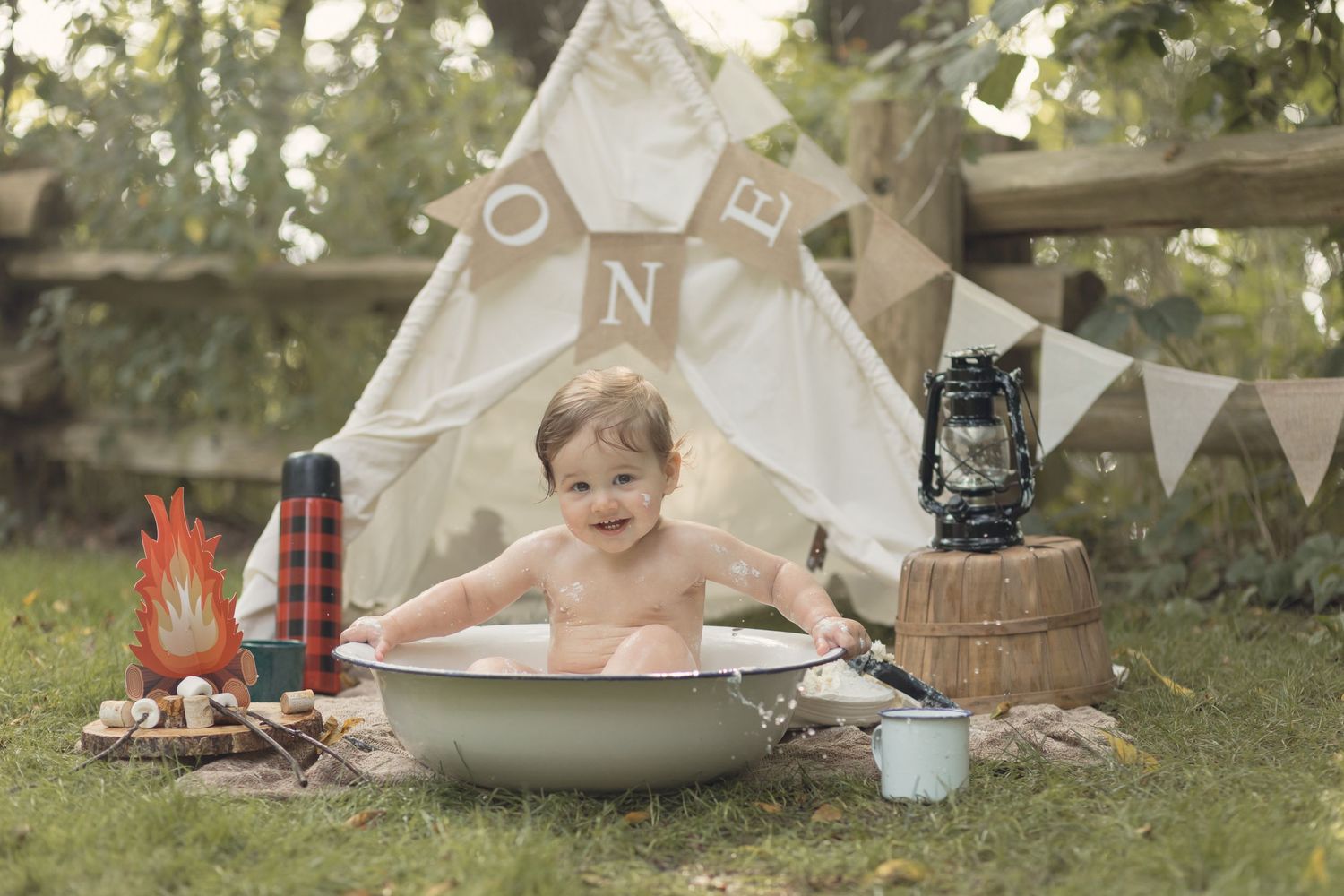 A child plays in a tin bath outside with a vintage camping setup featuring a teepee tent, lantern and plaid blanket.