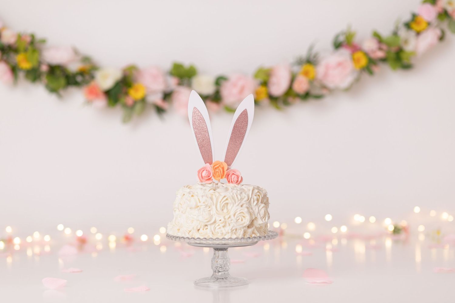 A white frosted cake with pink bunny ears on a cake stand, decorated with floral garland and twinkle lights.
