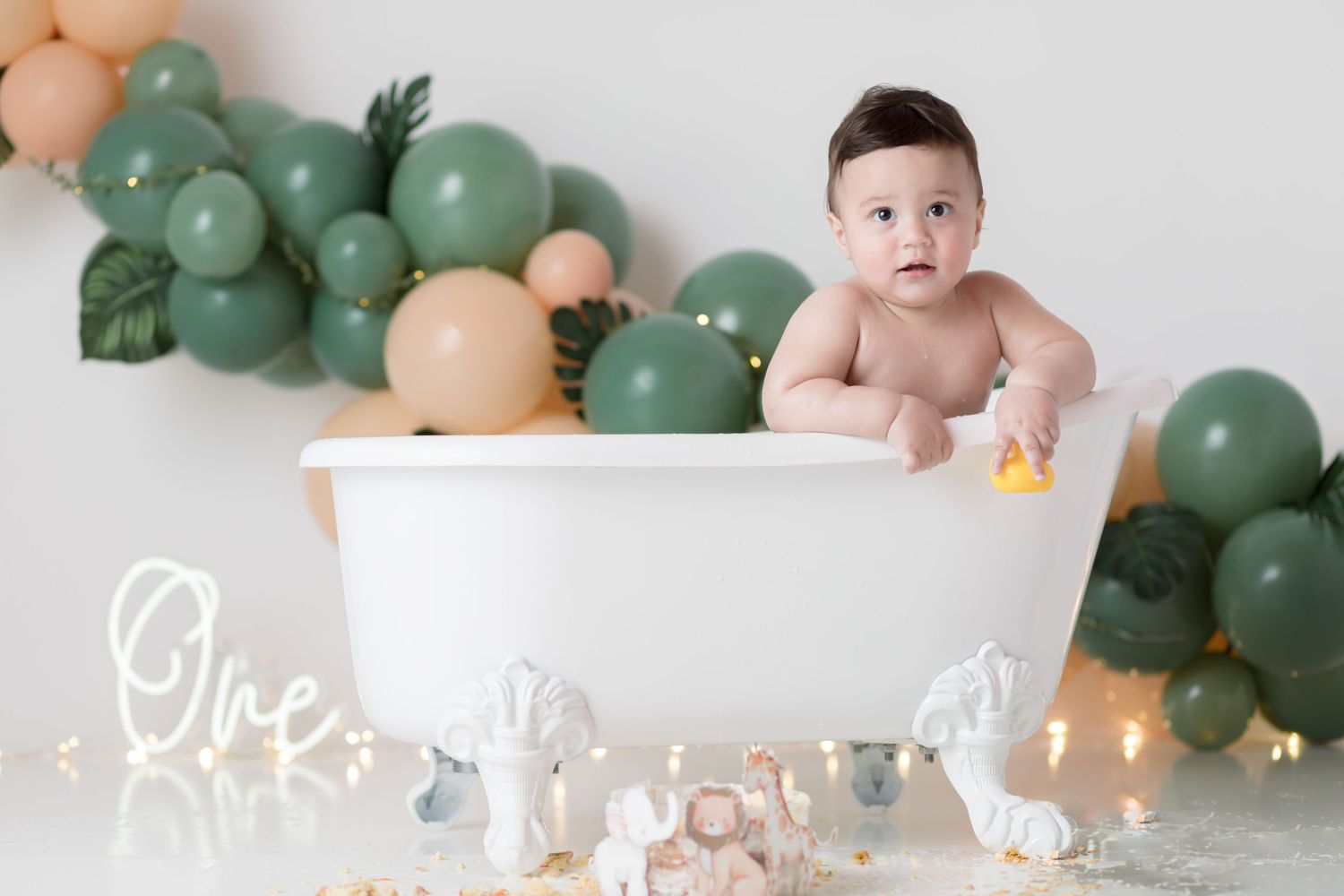 Baby sits in white bathtub with green and peach balloon arch and string lights in background.