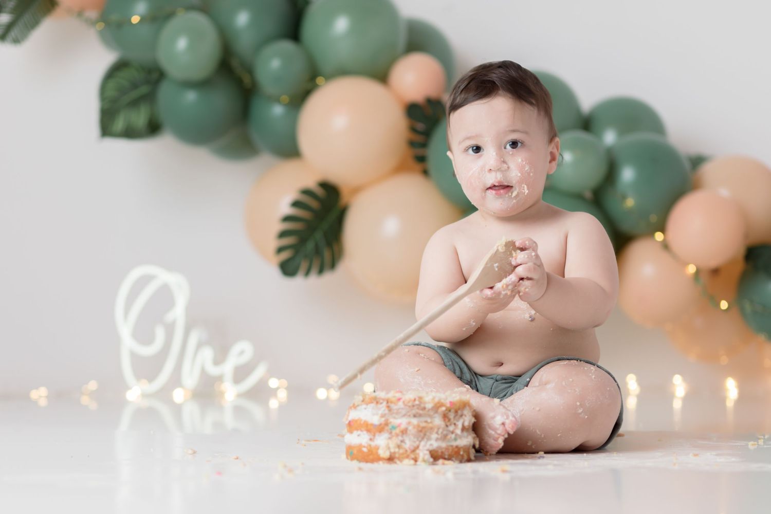 Baby sits with smash cake against decorative balloon arch with greenery and string lights.