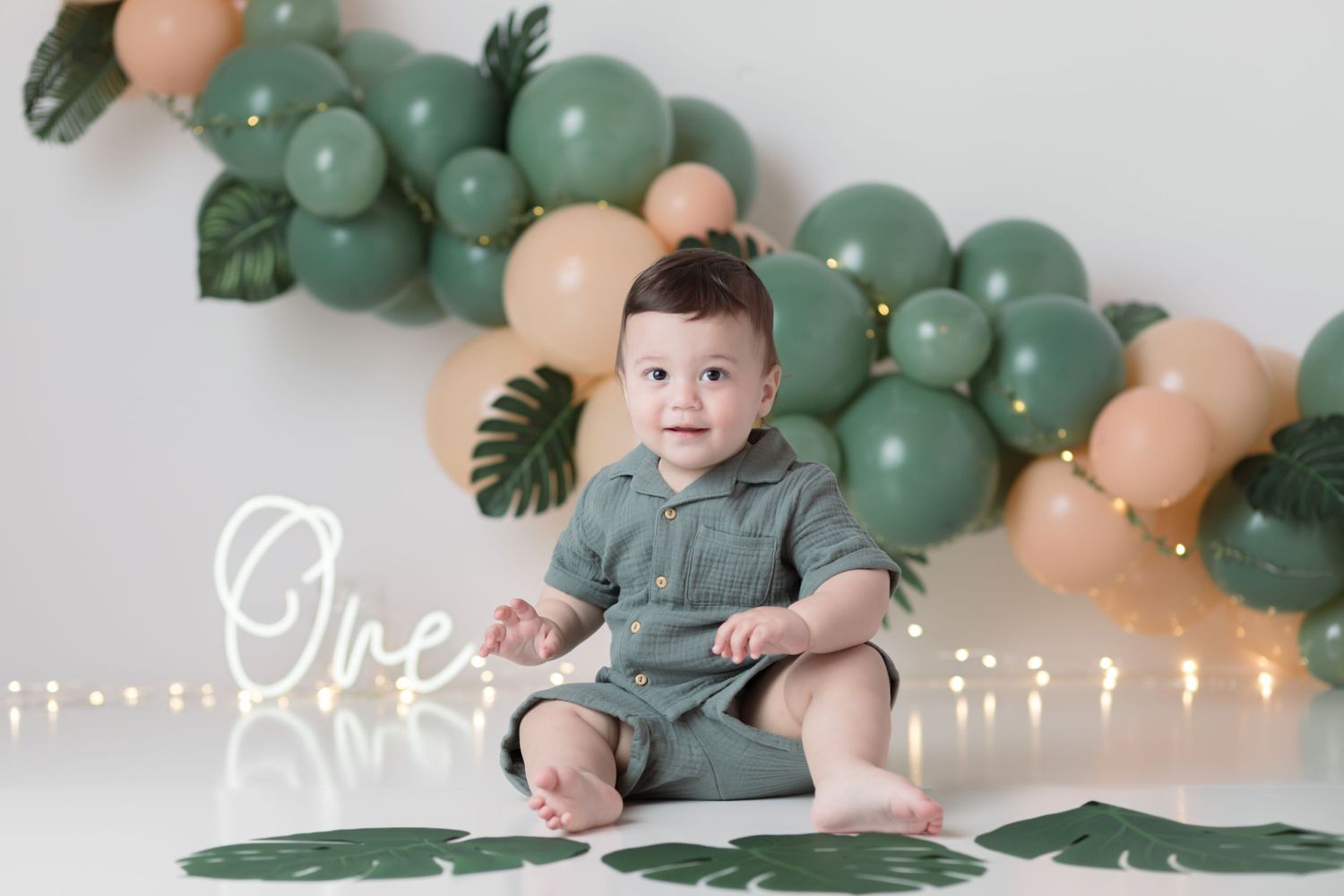 Baby in green outfit sits with cake against green and peach balloon garland backdrop.
