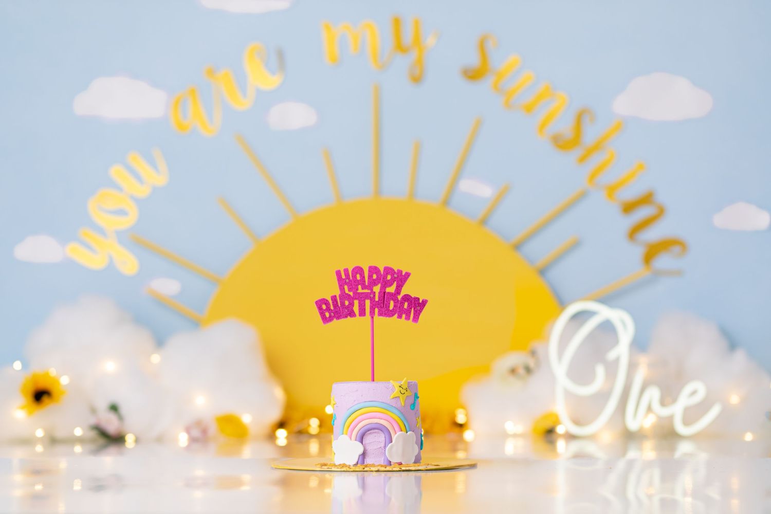 Birthday decoration with yellow sun backdrop, pink cake and cursive text against blue background.