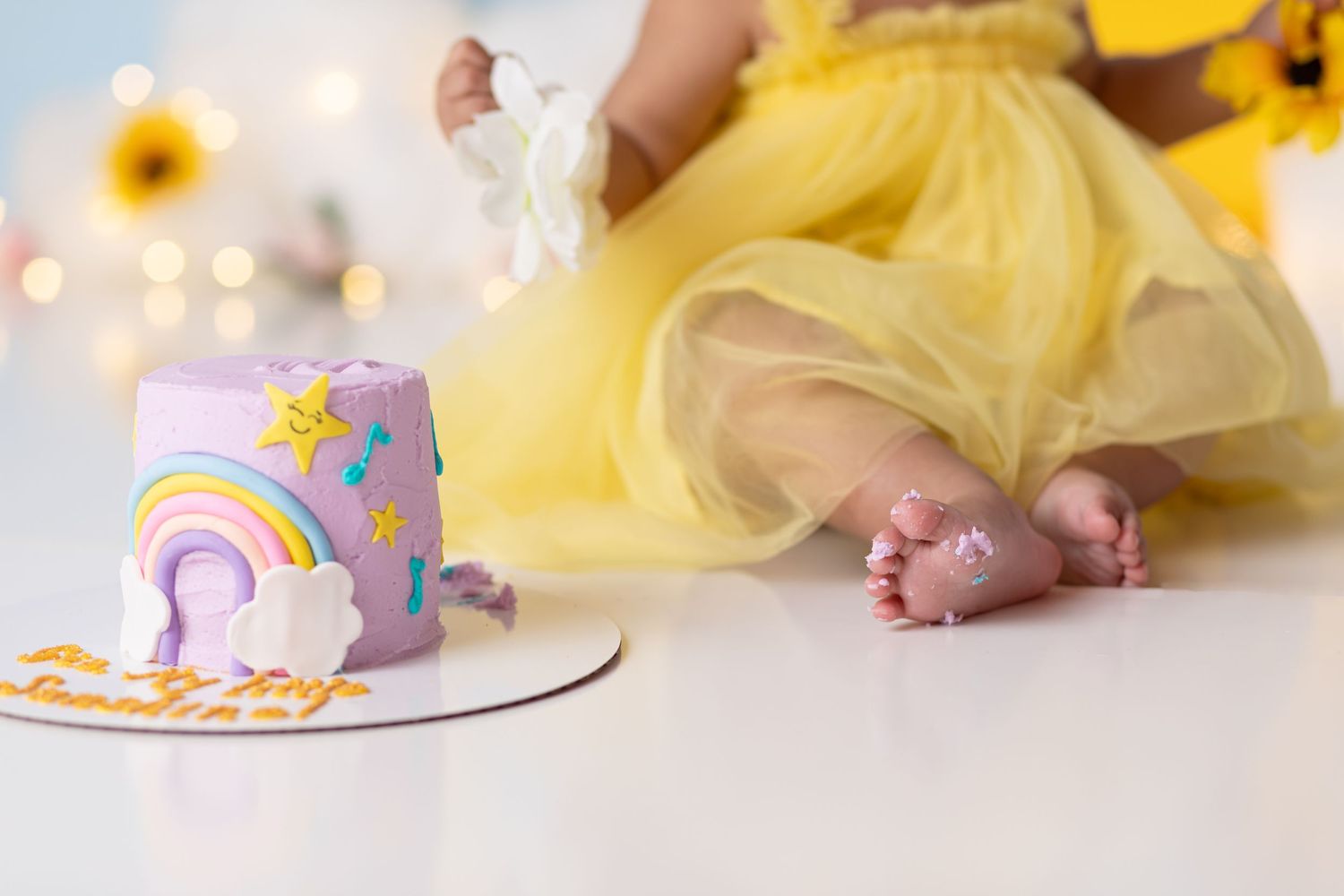 Small bare feet peek out from under a yellow tulle skirt next to a pastel rainbow birthday cake.