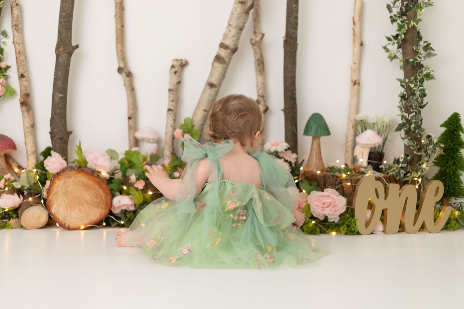 A small child in a mint green fairy dress sits in a whimsical woodland-themed set with birch logs and flowers.