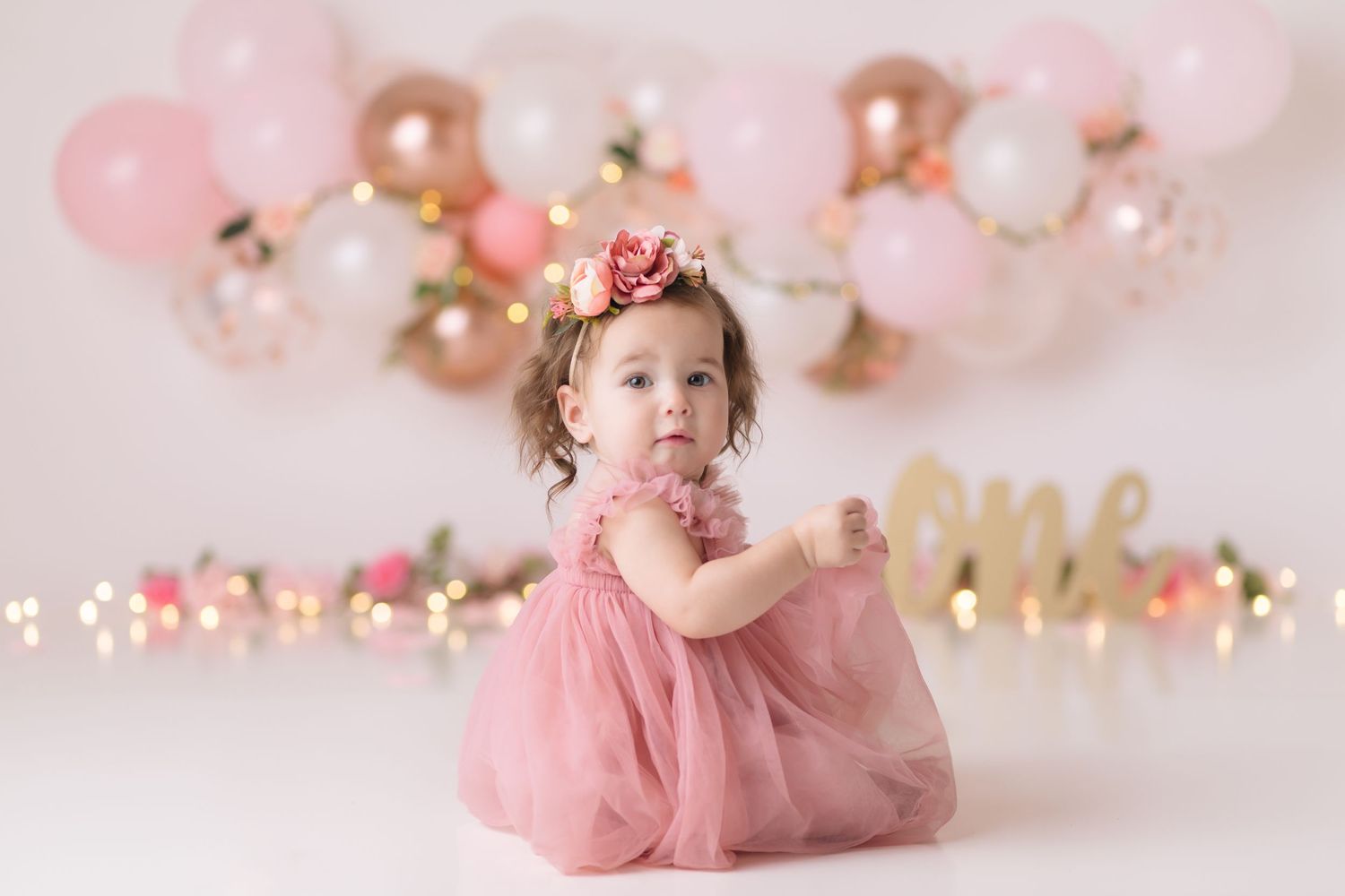Pink and gold themed first birthday photo setup with balloons and decorations against soft pink background.