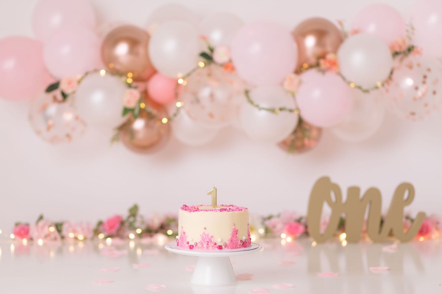 Pink and gold balloon garland with birthday cake setup against soft pink background.