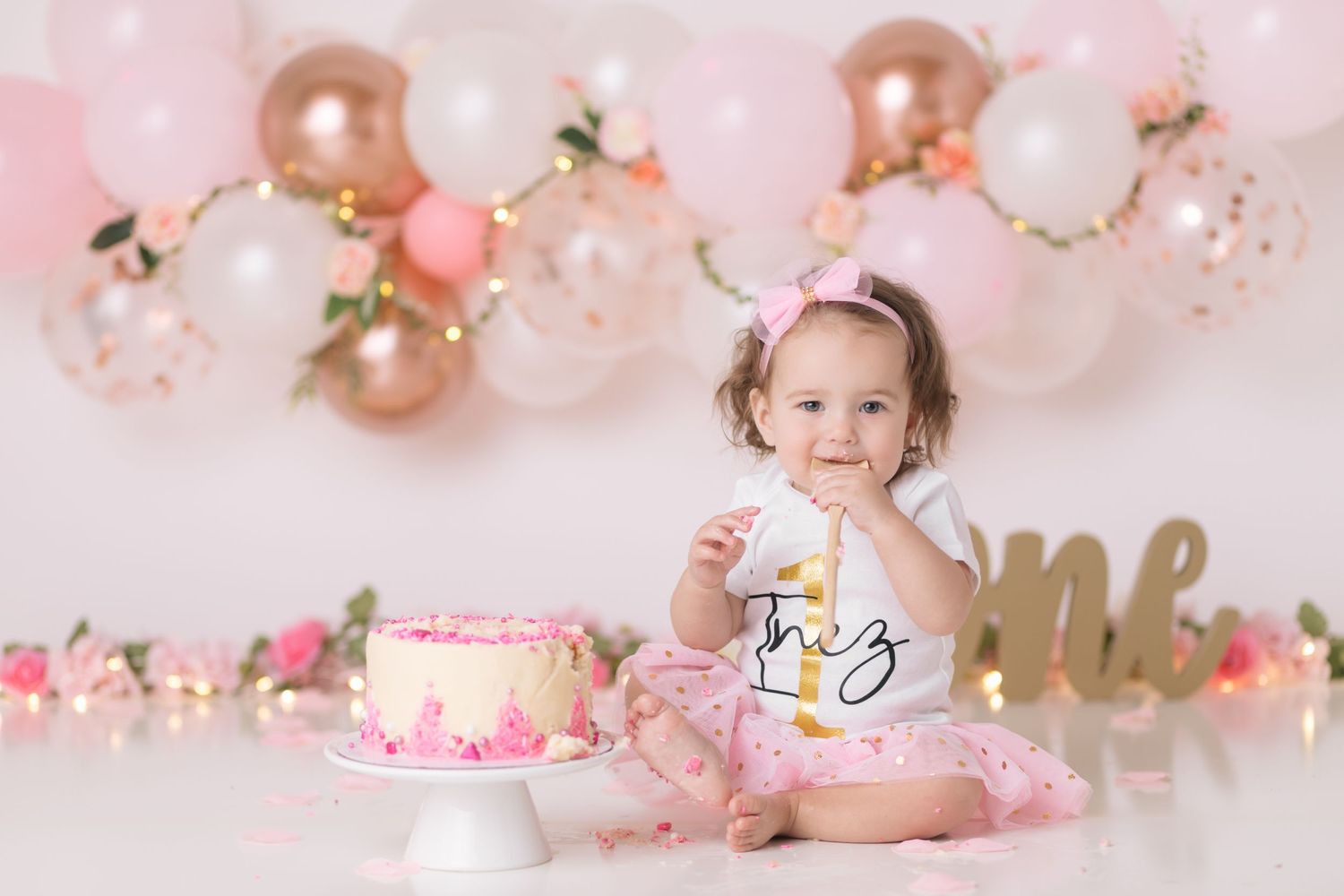 First birthday celebration setup with pink and gold balloons, floral decorations, and a pink cake against white backdrop.