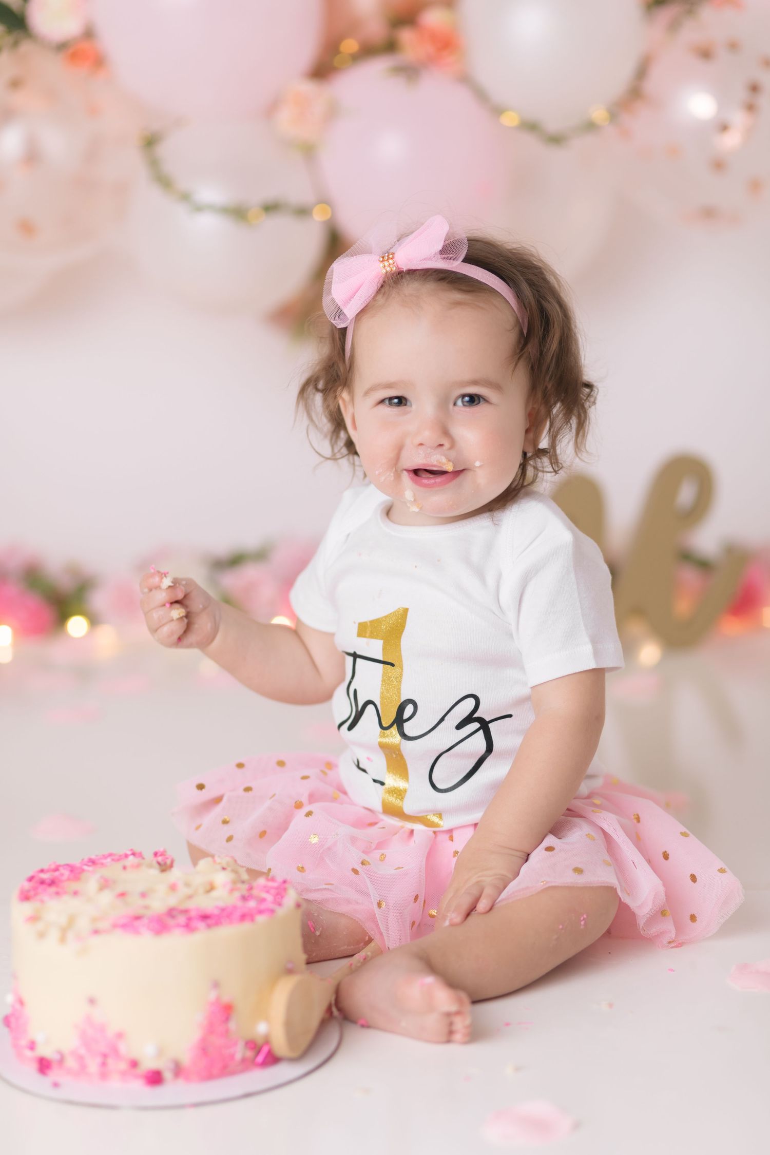 A first birthday celebration scene with pink balloons, tulle skirt, and birthday cake decorated in pink accents.