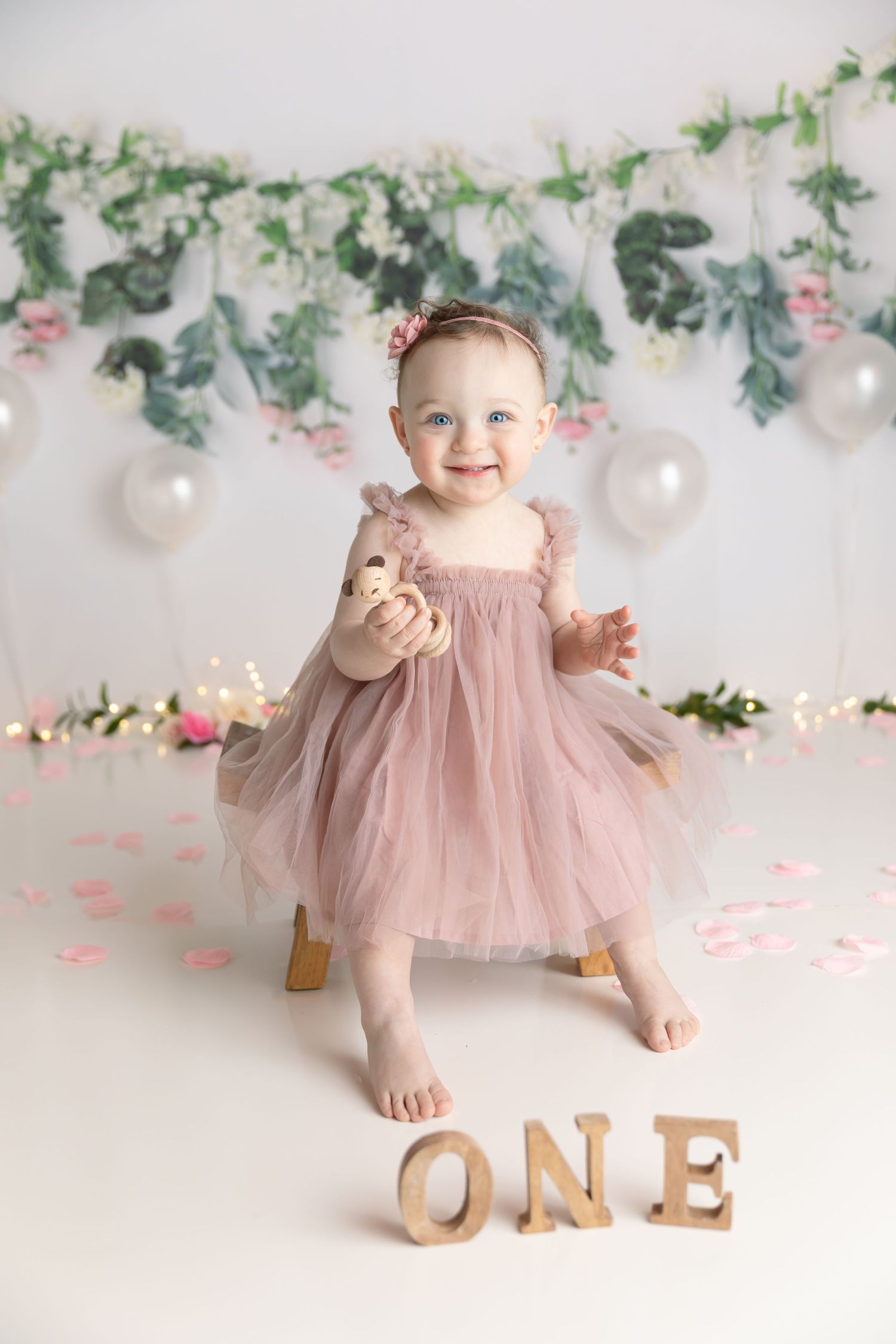Baby in pink tulle dress stands by wooden ONE letters with floral garland backdrop.