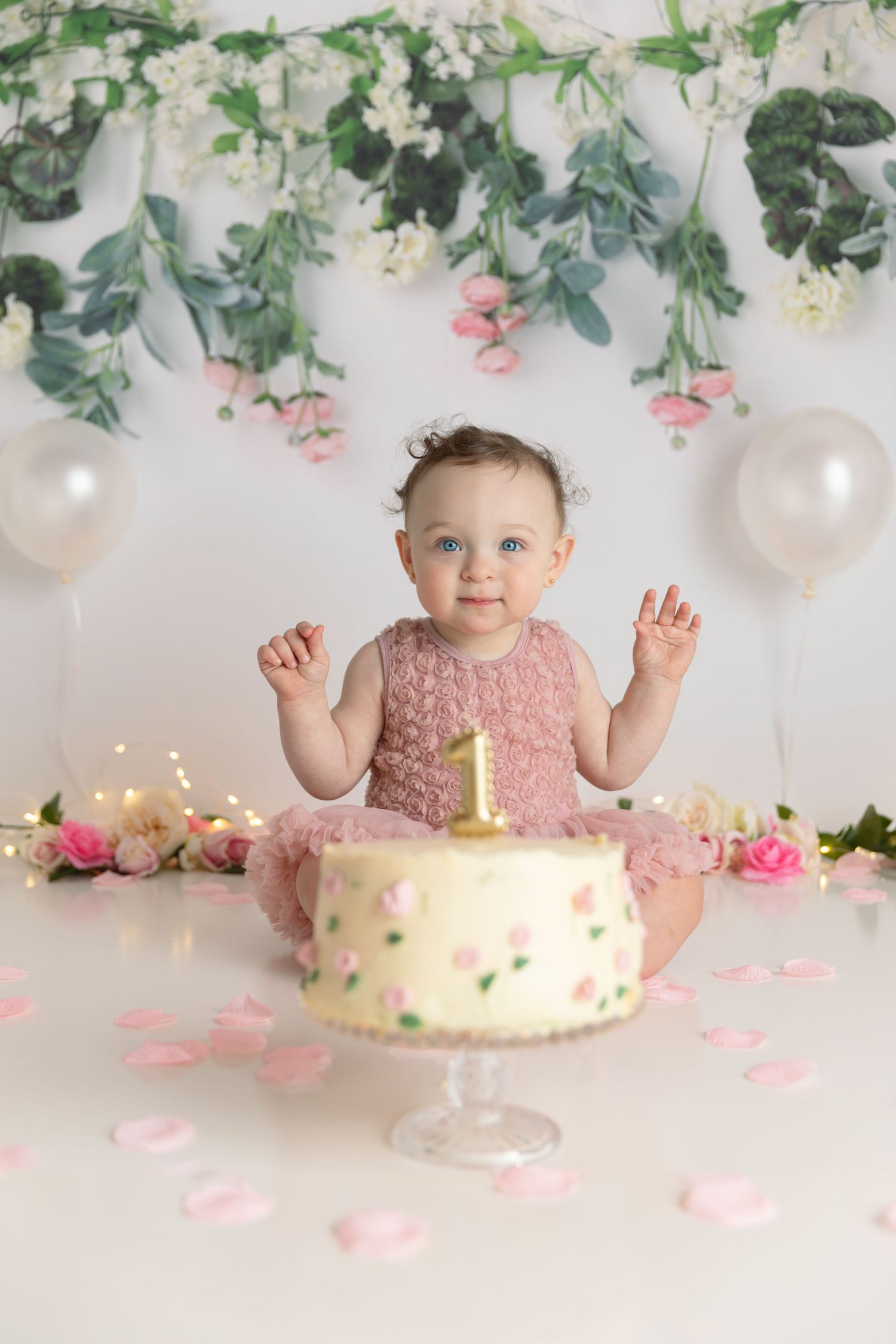 Baby in pink dress sits with birthday cake against floral backdrop with white balloons.