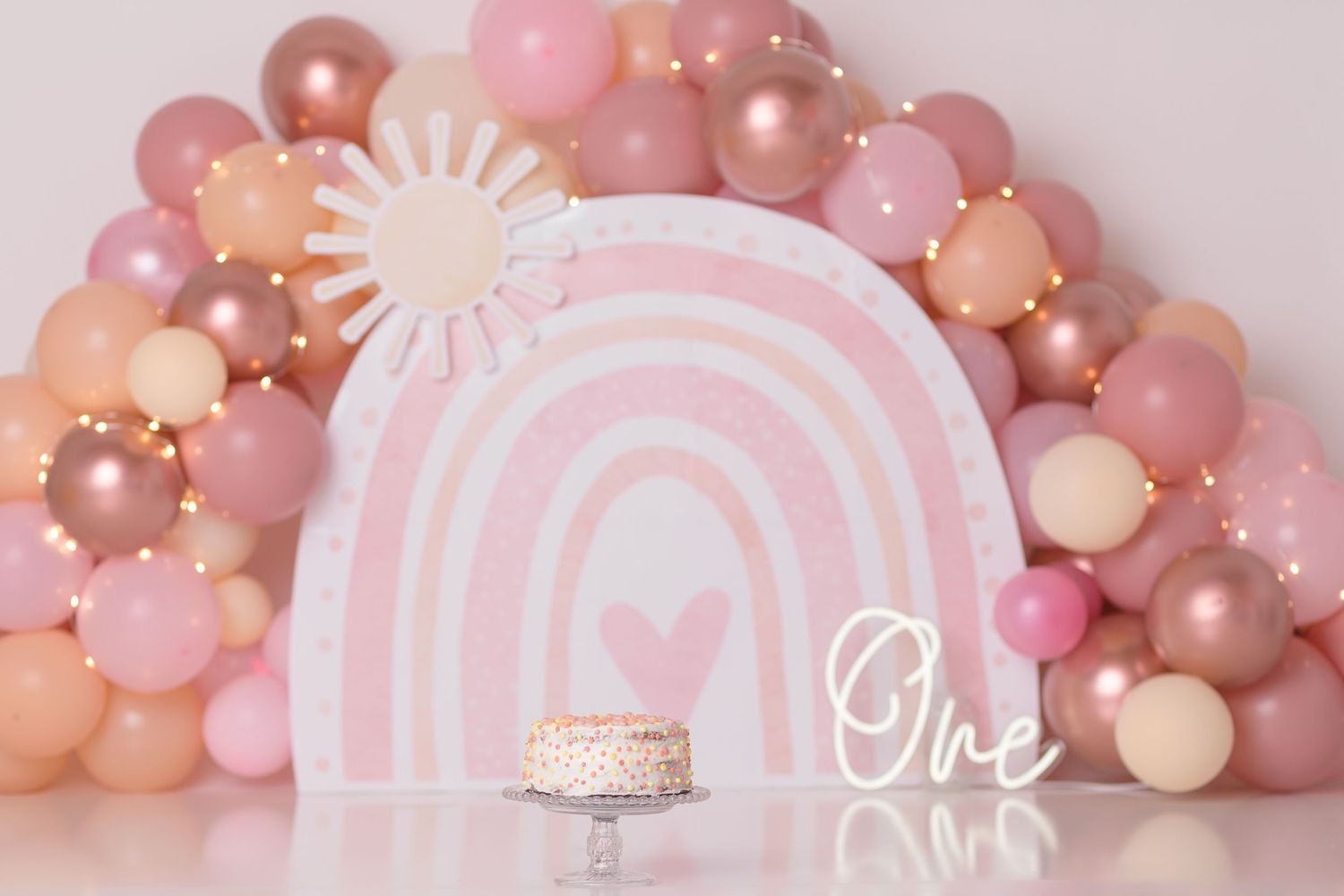 Pink and peach balloon arch with rainbow design backdrop for first birthday celebration.