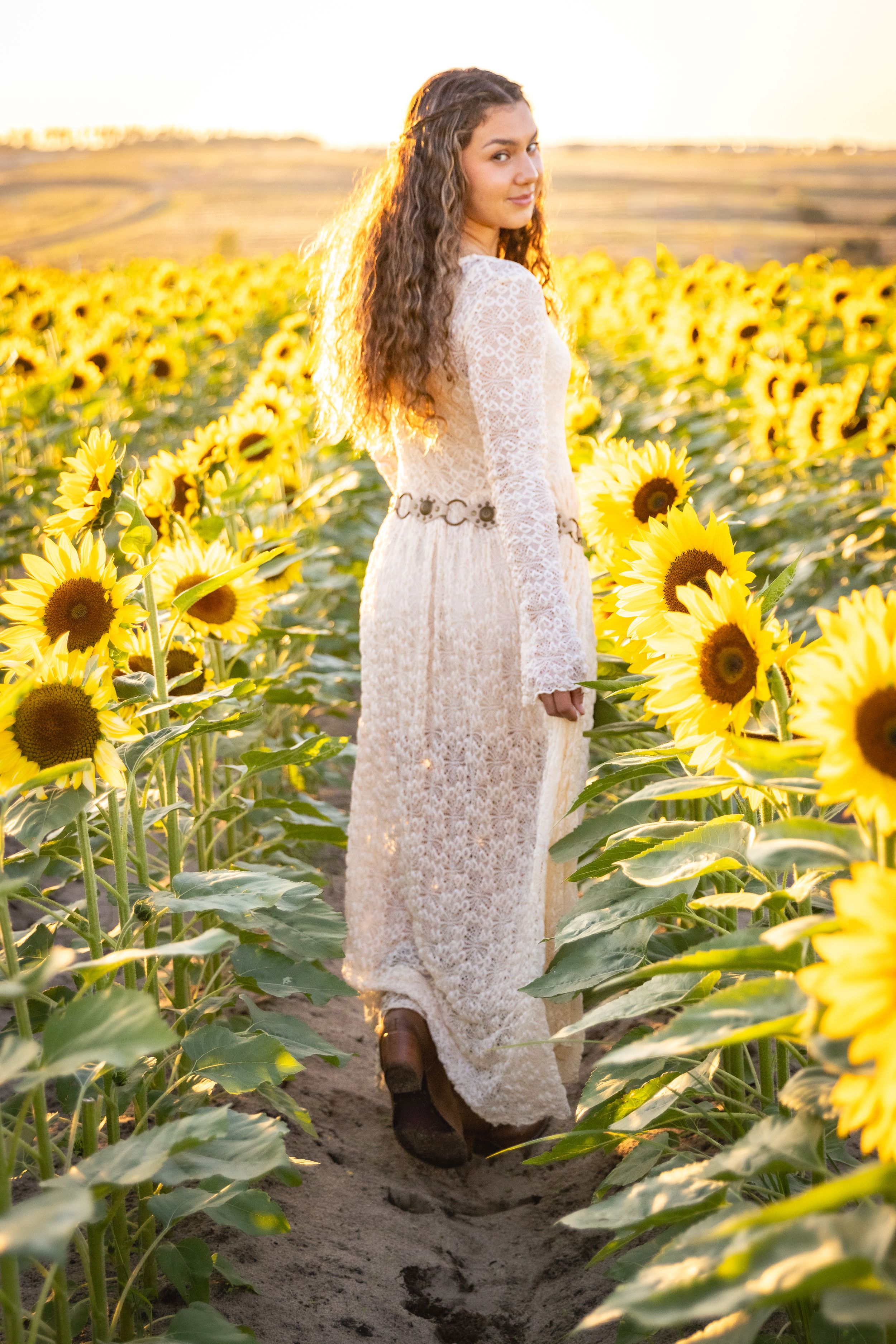 Young woman in white lace dress standing in sunflower field at golden hour, smiling over shoulder.