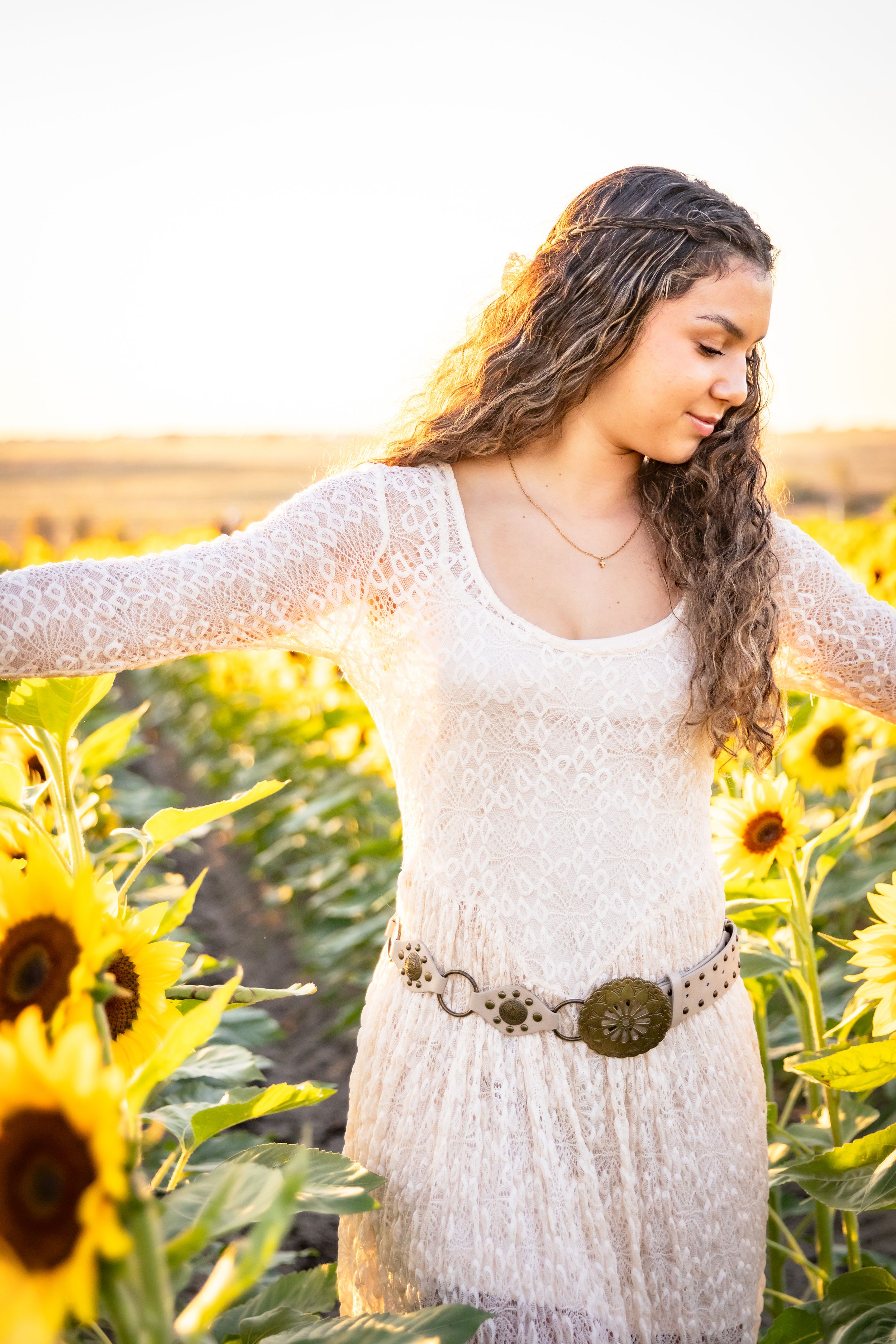 Beautiful girl in white lace dress with belt posing in sunflower field at sunset.
