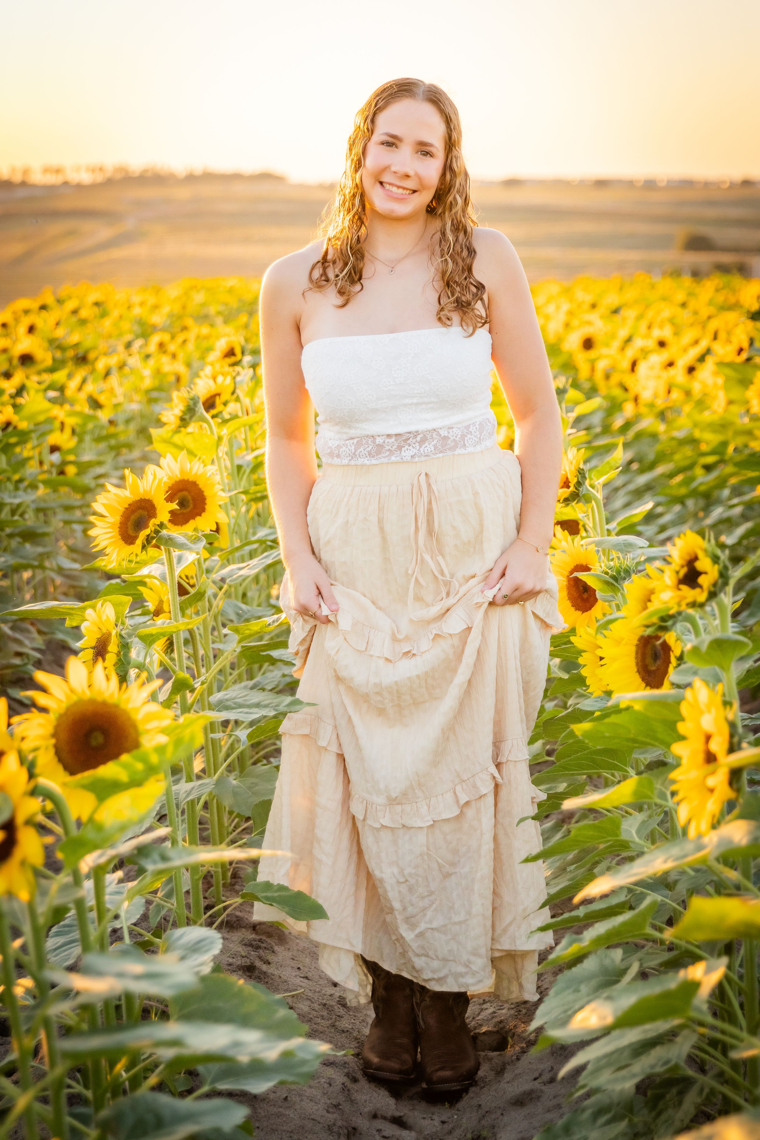 Young woman in white top and beige ruffled skirt smiling in a sunflower field at golden hour.