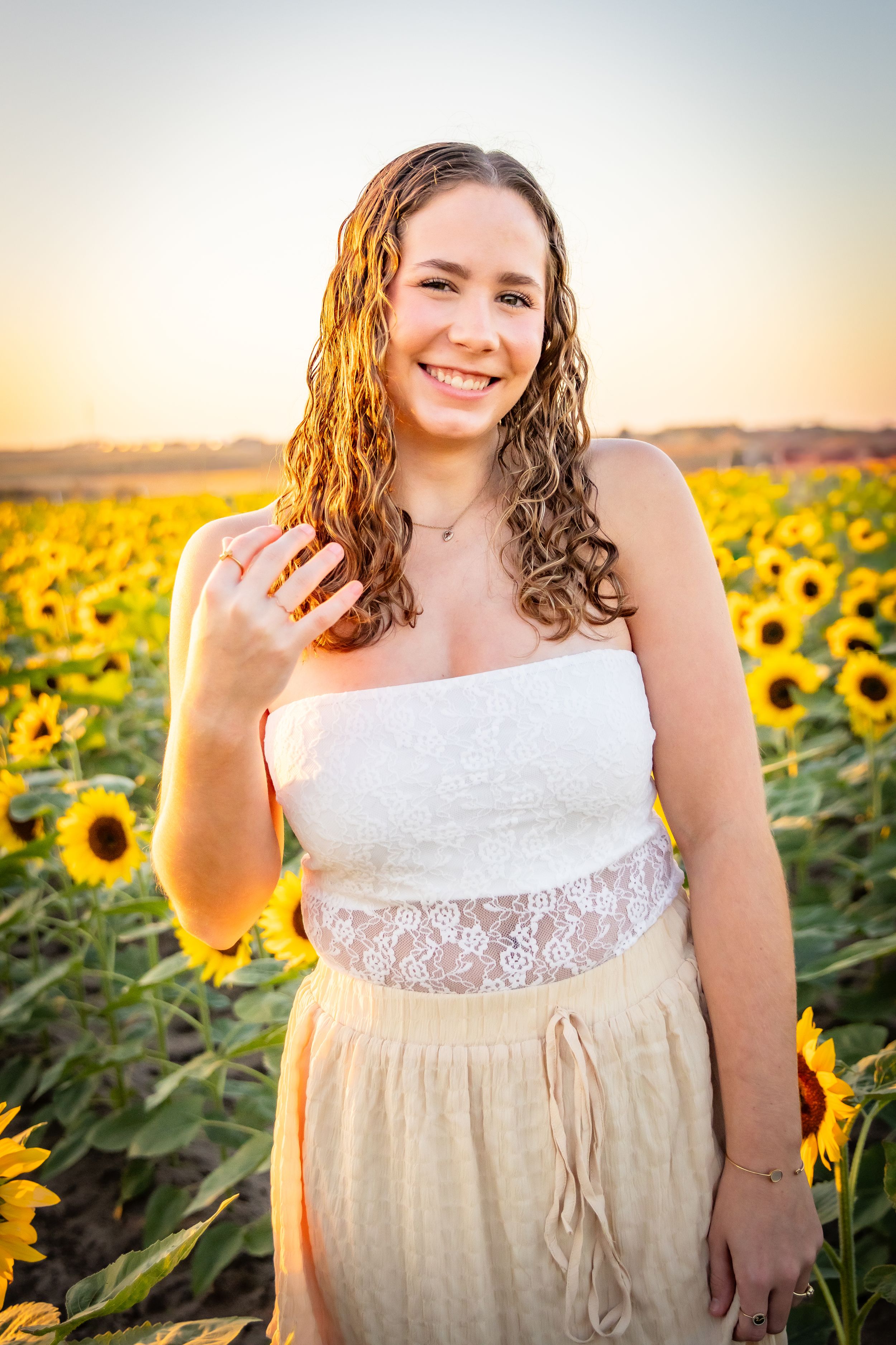 Smiling young woman in white lace top and yellow skirt posing in a vibrant sunflower field at golden hour.