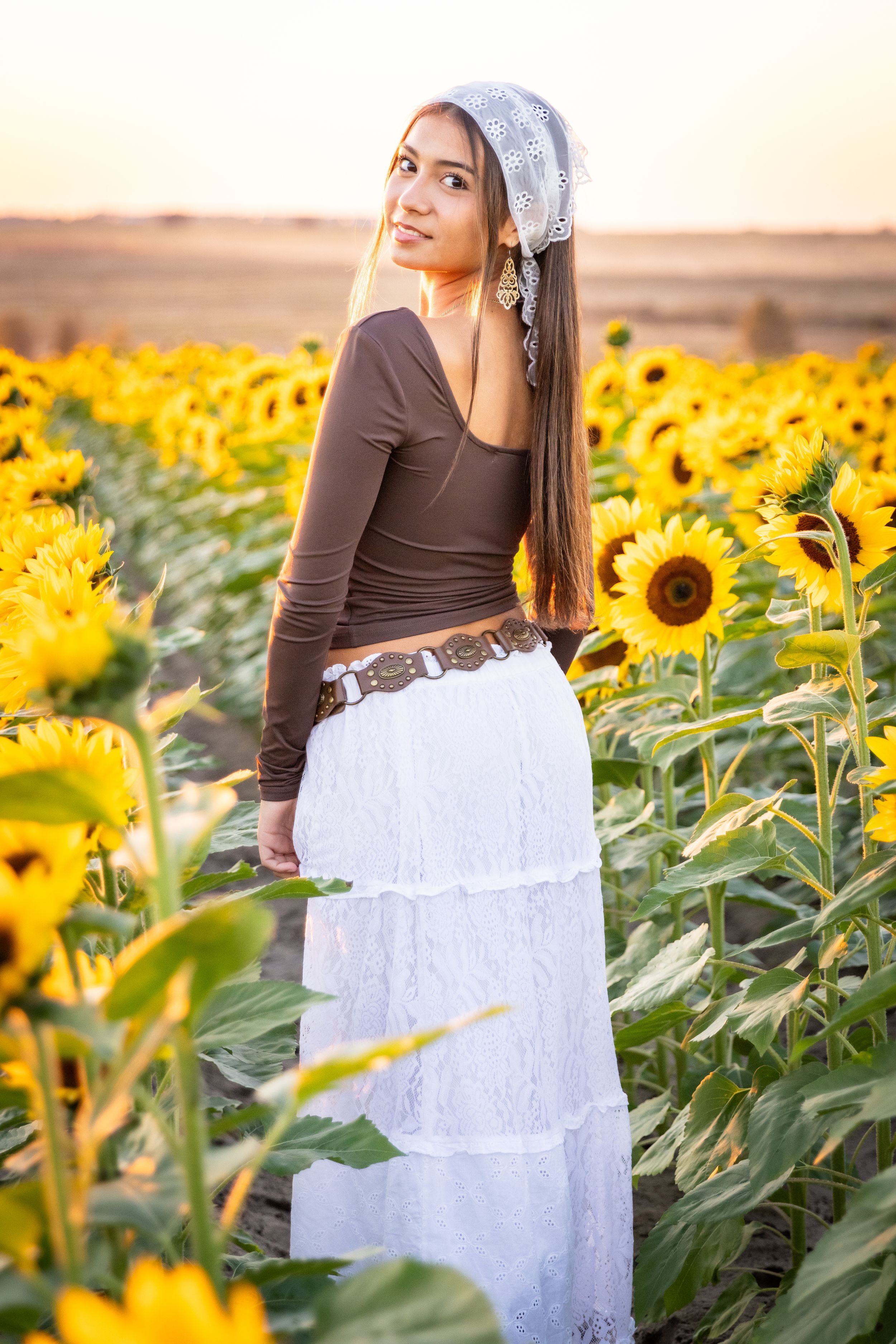 Young woman in white skirt and brown top poses gracefully in a vibrant sunflower field at golden hour.