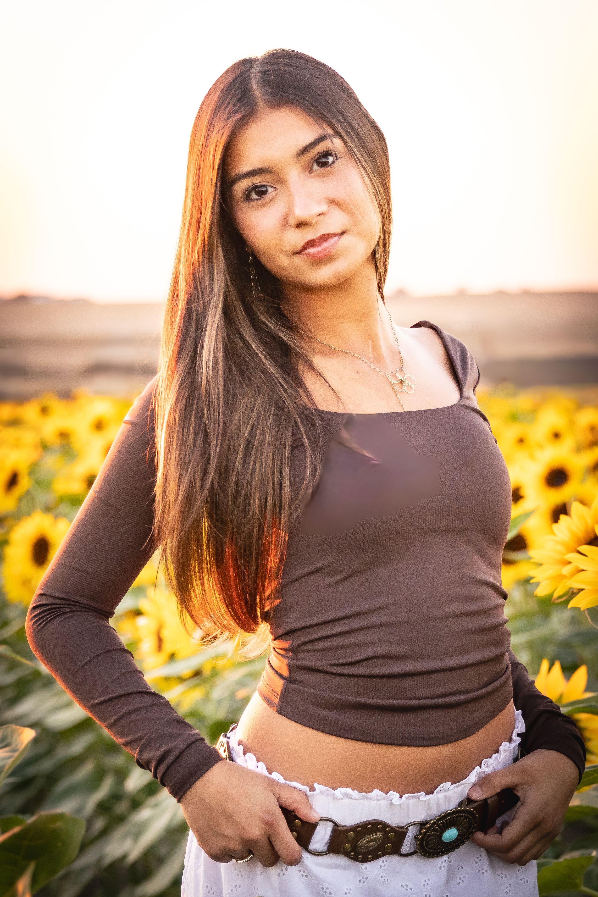 Young woman in brown crop top and white skirt poses in a vibrant sunflower field at golden hour sunset.