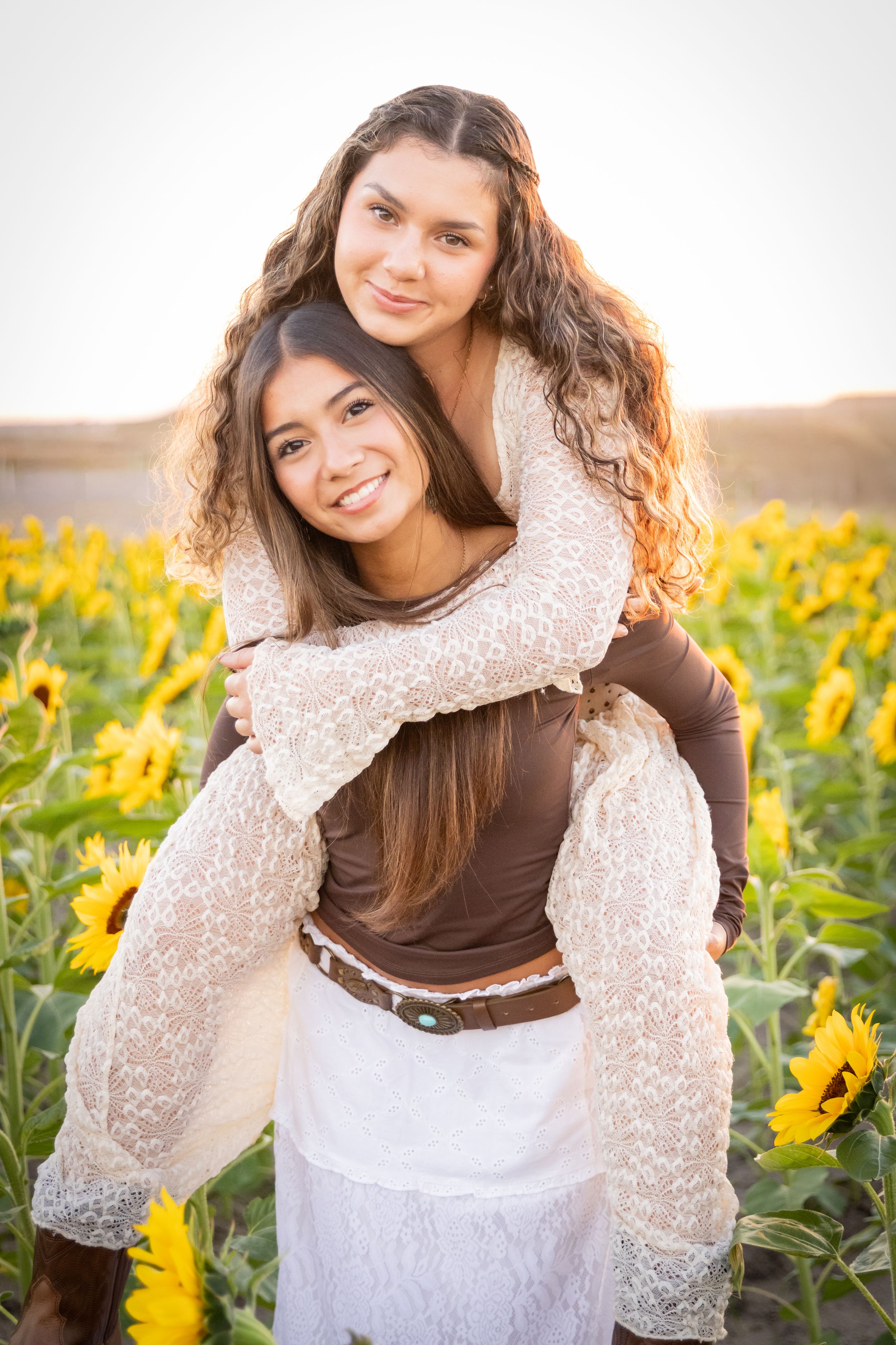 Two young women posing in a sunflower field, one giving the other a piggyback ride at golden hour.