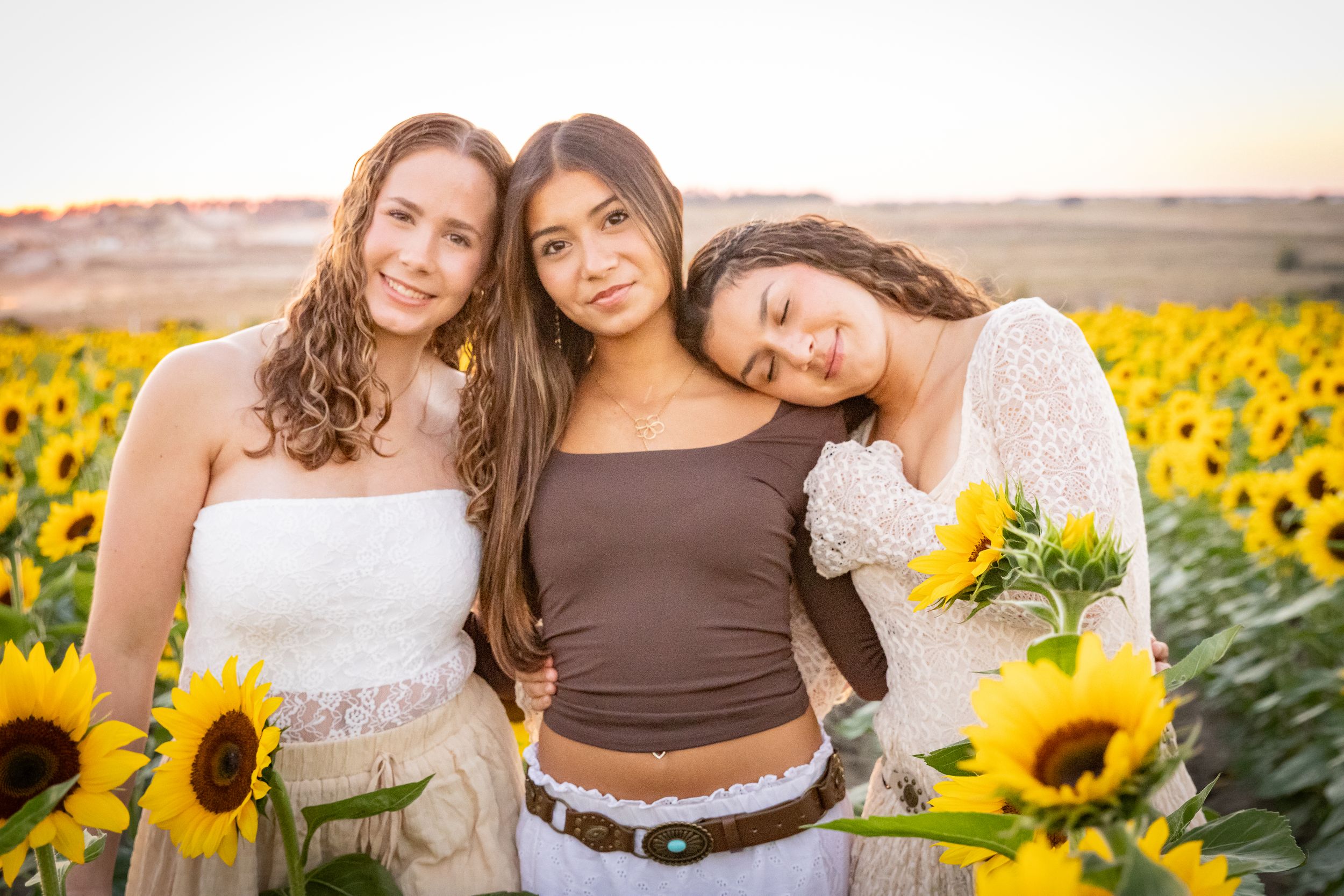 Three young women posing together in a sunflower field at sunset, holding yellow sunflowers.