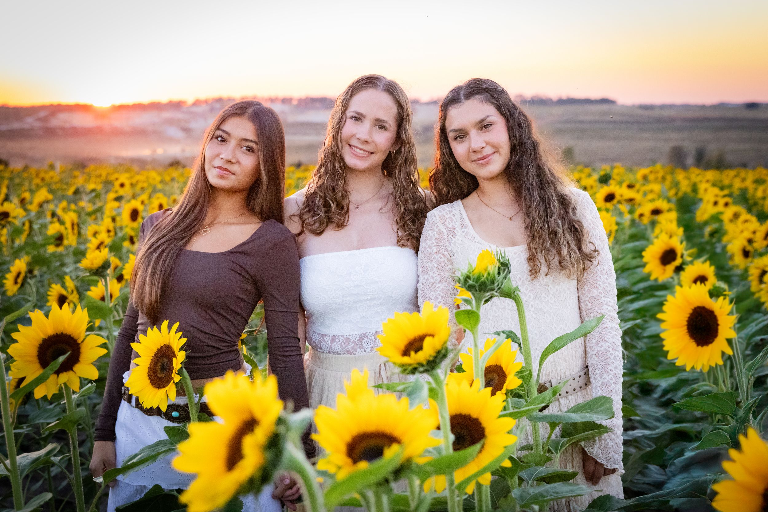 Three young women smiling in a sunflower field at golden hour sunset.