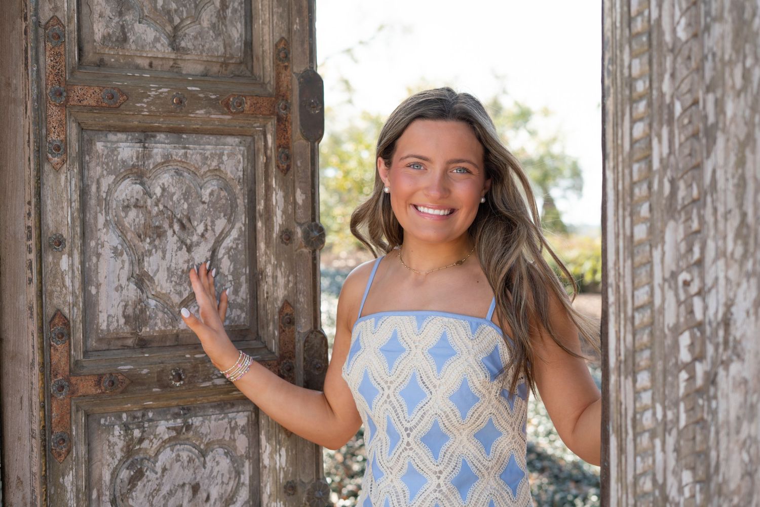 Smiling young woman in blue sundress posing by ornate rustic wooden doors outdoors.