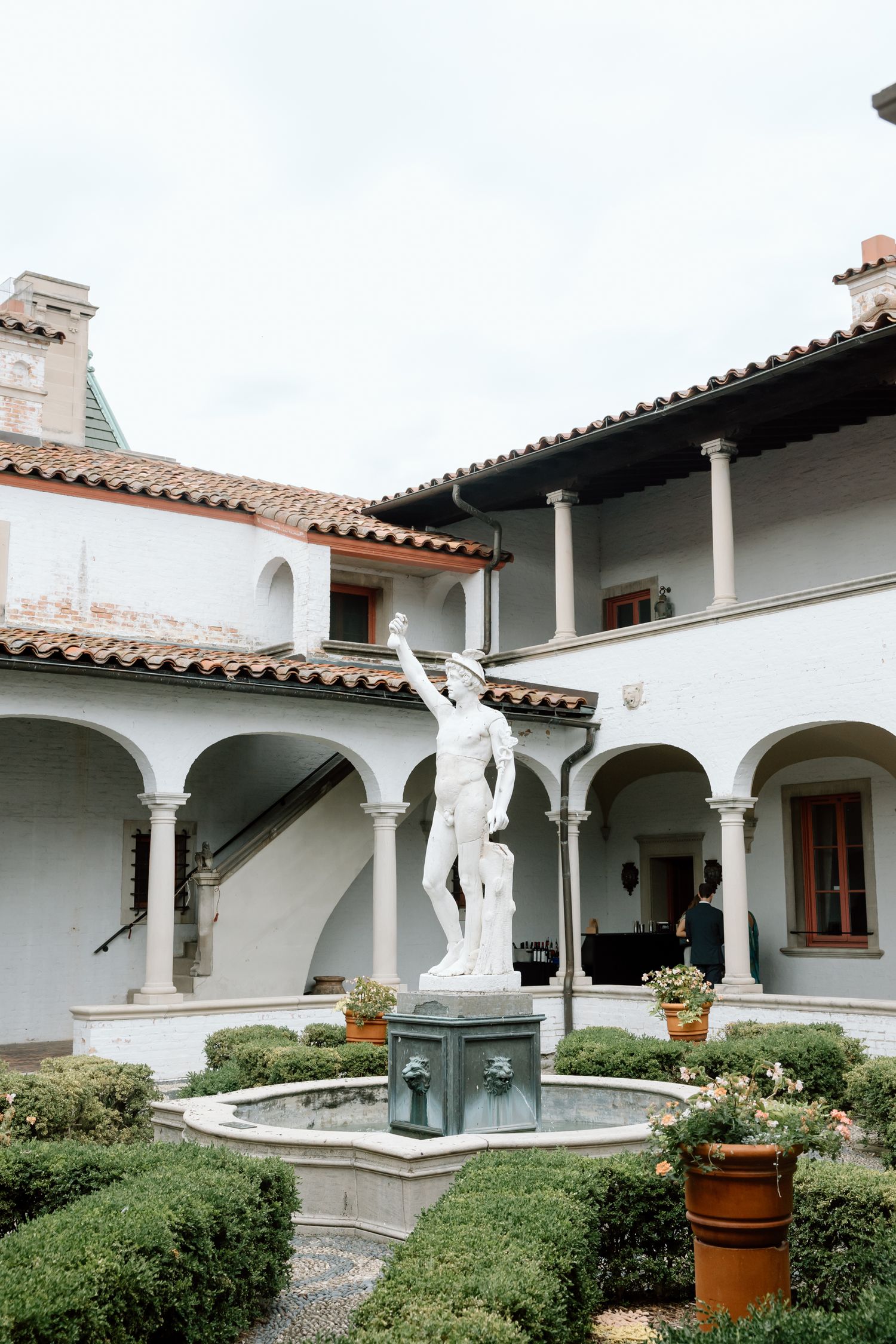 White classical statue fountain in Italian Renaissance courtyard with arched colonnade, terracotta roof, and manicured hedges.