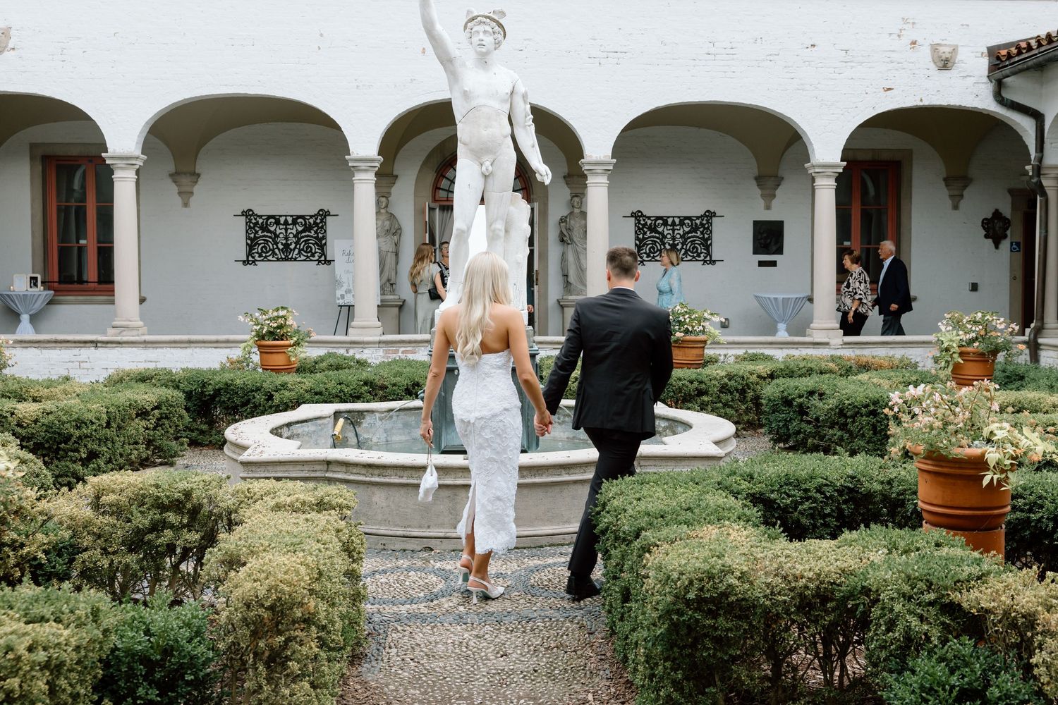 Couple walks hand-in-hand toward elegant garden fountain with classical statue at a luxurious Italian villa wedding.