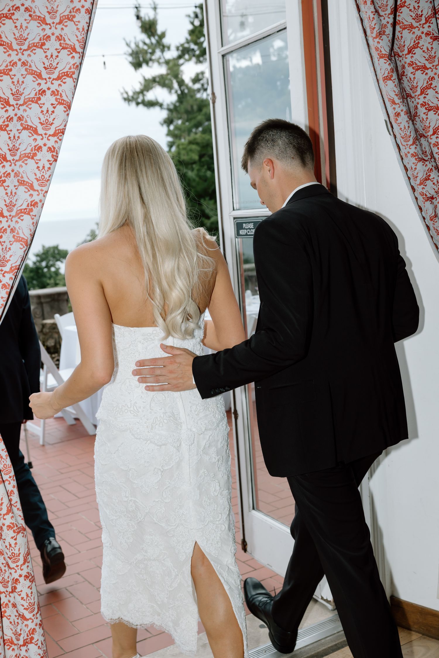 Bride in white lace dress and groom in black suit walking through decorated doorway to outdoor wedding venue.