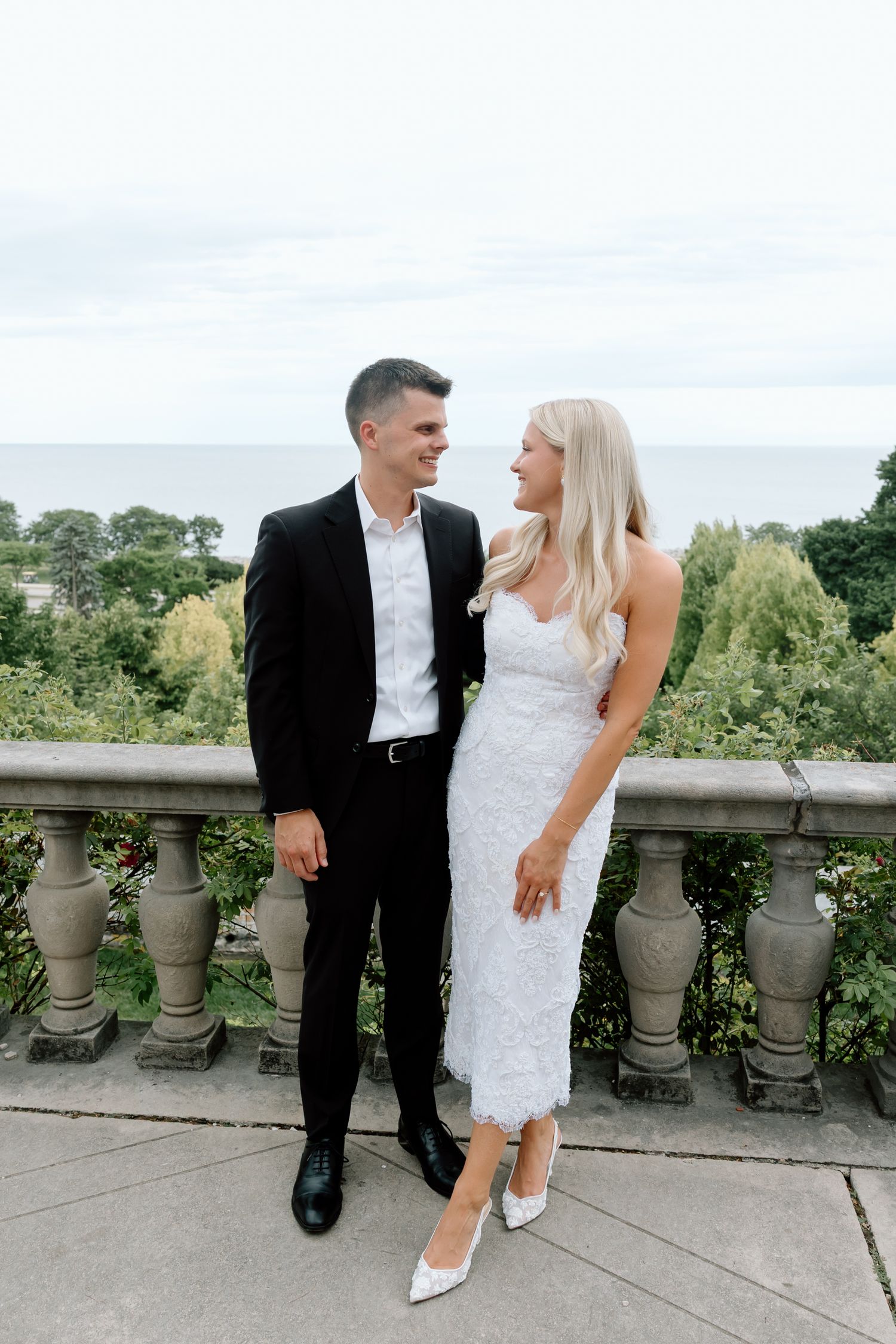 Couple in wedding attire smiling at each other on a stone balcony overlooking lush greenery and the ocean.