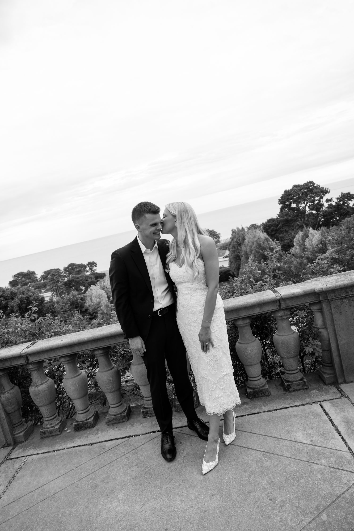 Black and white photo of a couple in wedding attire sharing a romantic moment on an elegant stone balcony.