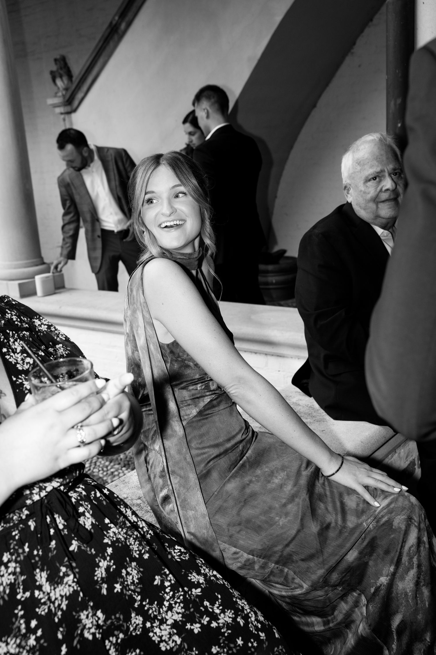 Black and white photo of a smiling young woman in elegant dress at a formal event with guests in background.