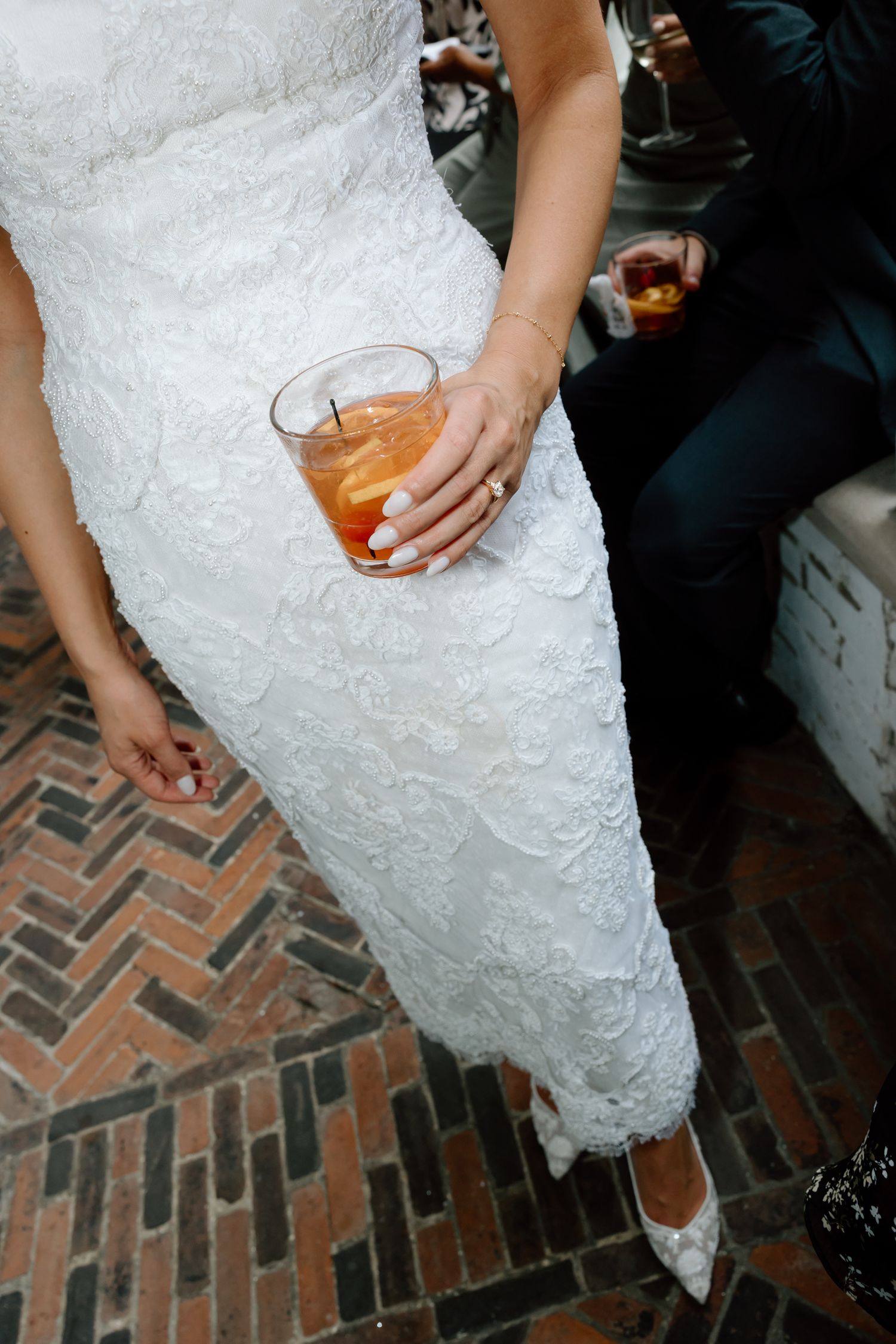 Bride in white lace dress holding an amber cocktail drink at a wedding reception on brick flooring.