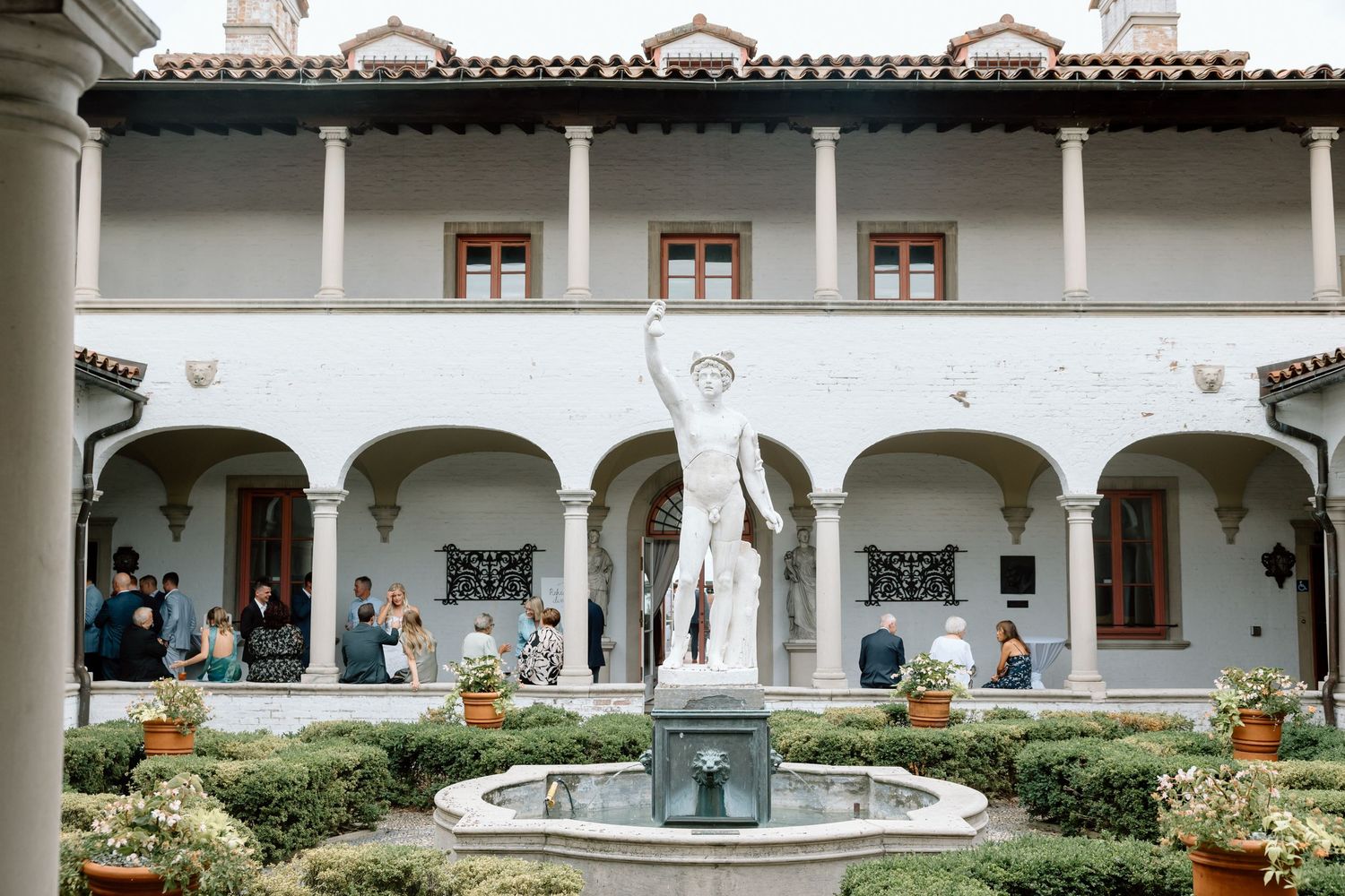 Elegant Italian villa courtyard with classical statue fountain, arched colonnade, and wedding guests.