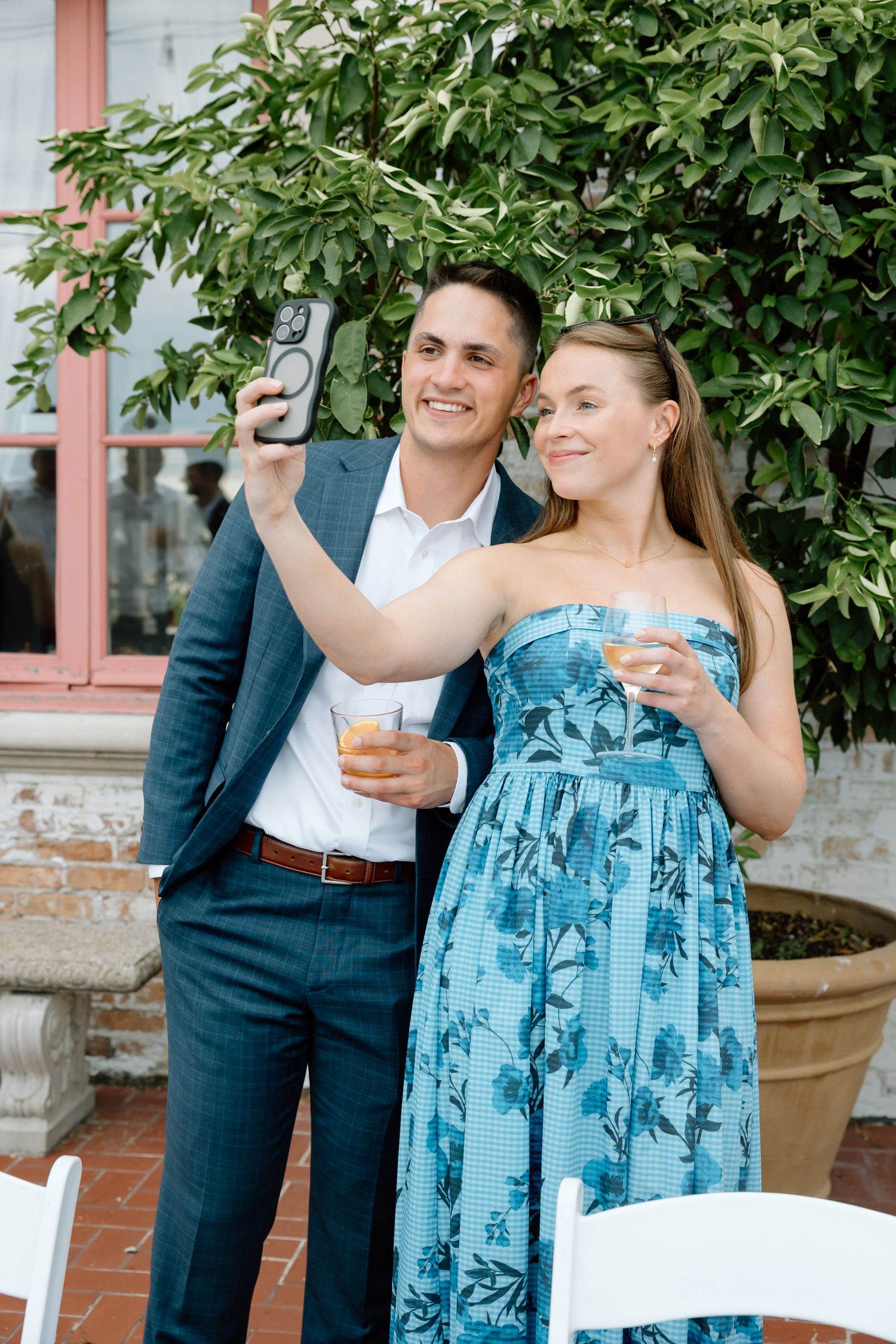 Couple taking a selfie outdoors at a garden party, holding cocktails, dressed in blue floral dress and navy suit.
