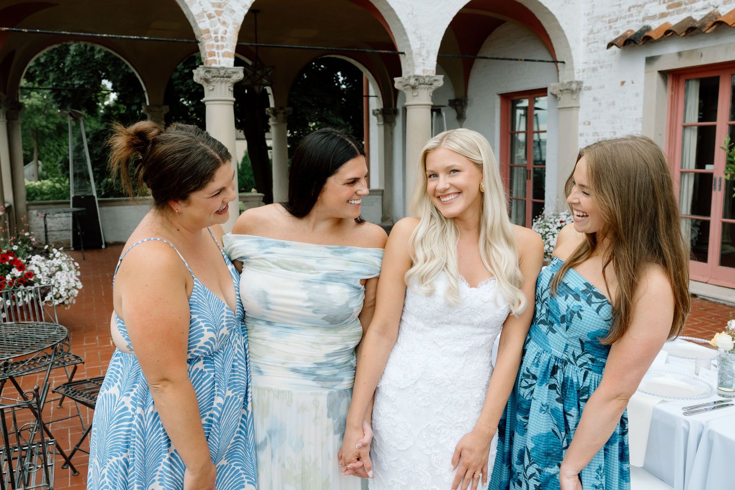 Bride in white gown laughing with three bridesmaids in blue floral dresses at outdoor wedding venue.