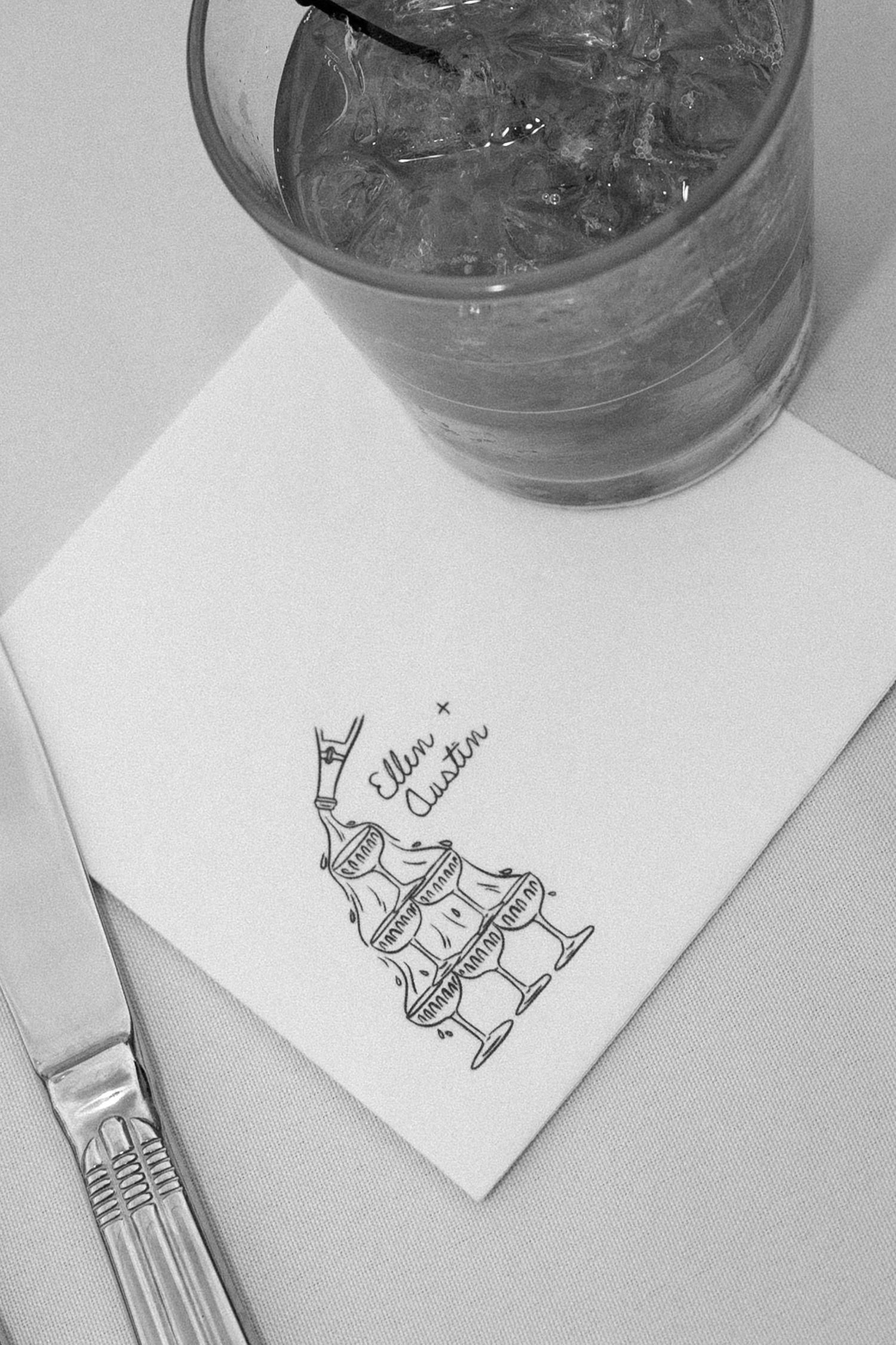 Custom wedding cocktail napkin printed with couple names Ellor and Auston beside a glass of iced drink and silverware.