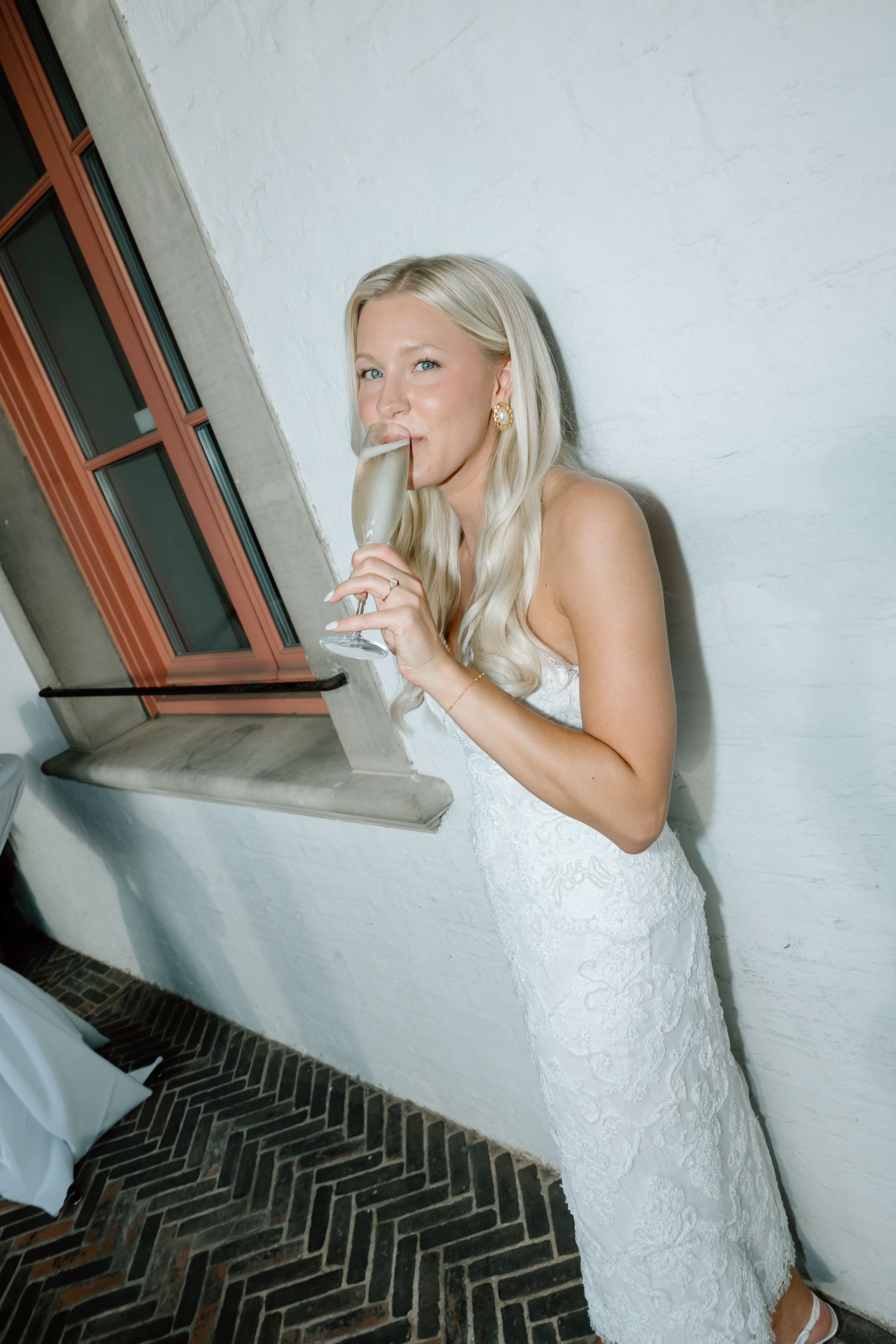 Blonde bride in white lace gown sipping champagne while leaning against a white brick wall near a window.