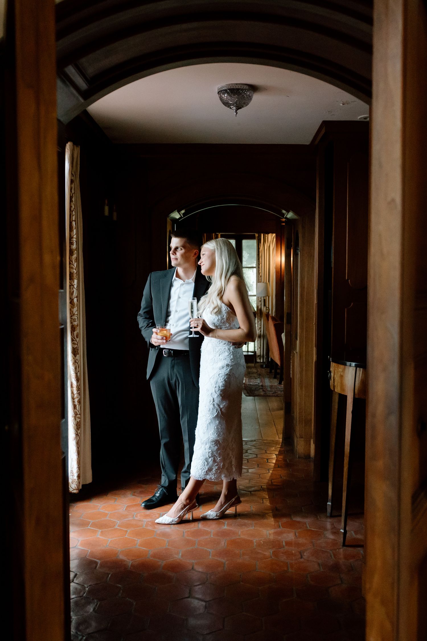 Bride and groom standing together in elegant dark wood hallway holding champagne glasses at wedding venue.