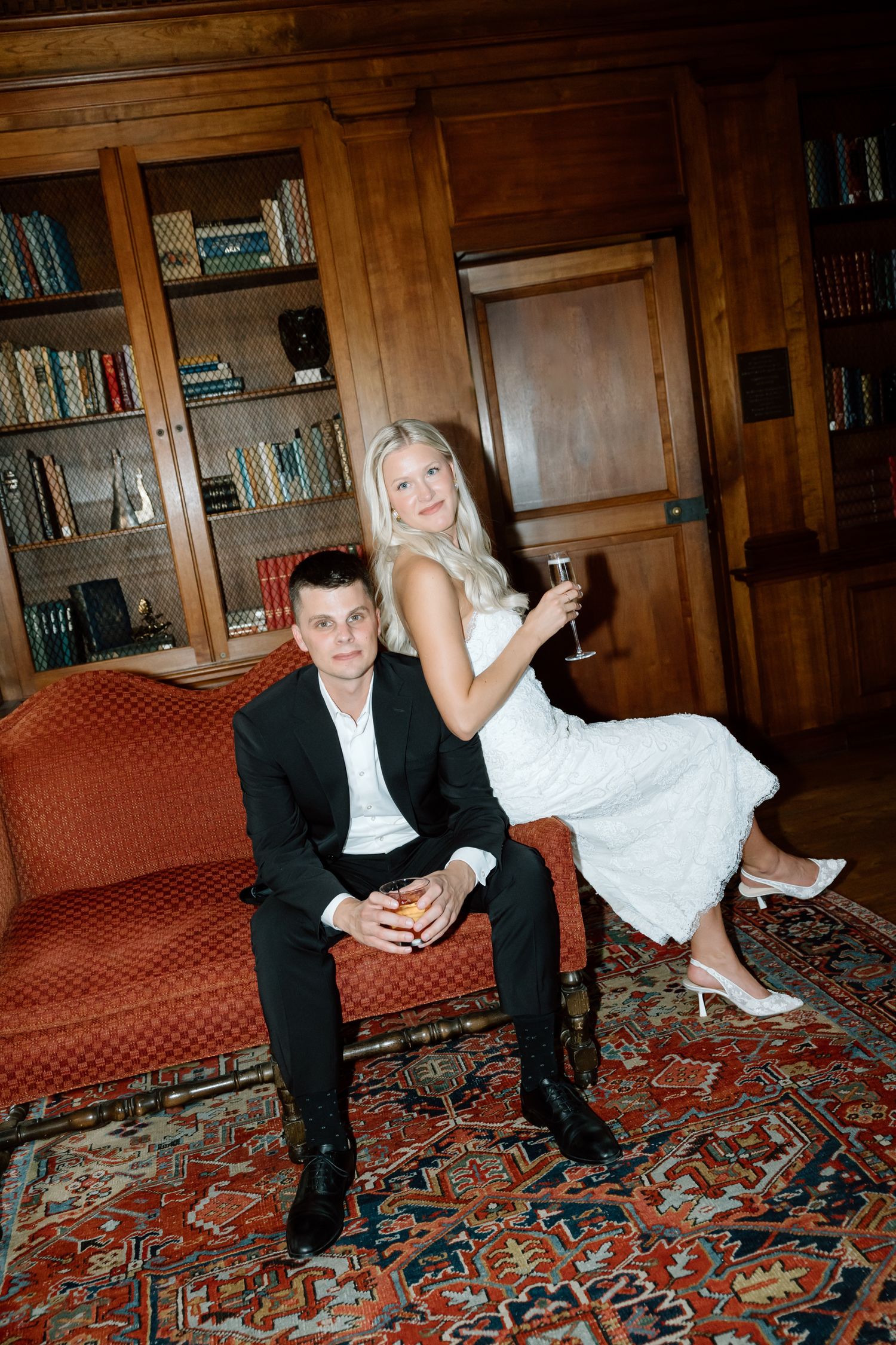 Elegant couple posing in a wood-paneled library, woman in white dress holding champagne, man seated on red sofa.