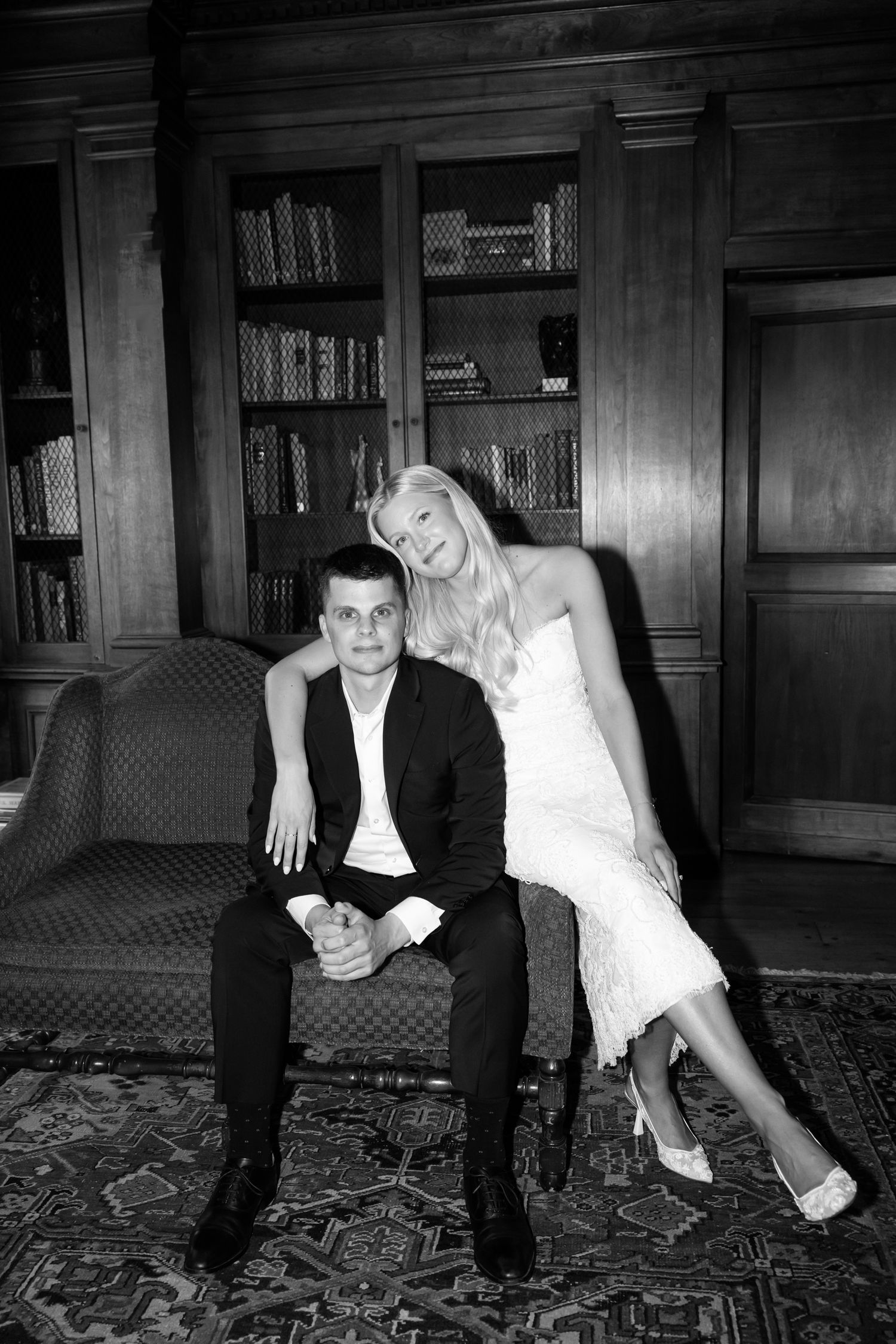 Black and white photo of bride and groom posing elegantly in a wood-paneled library with bookcase backdrop.