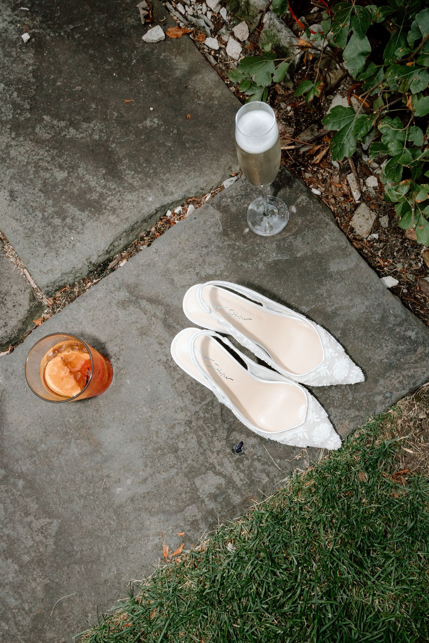 White bridal heels on stone pavement beside a champagne flute and cocktail at an outdoor wedding venue.