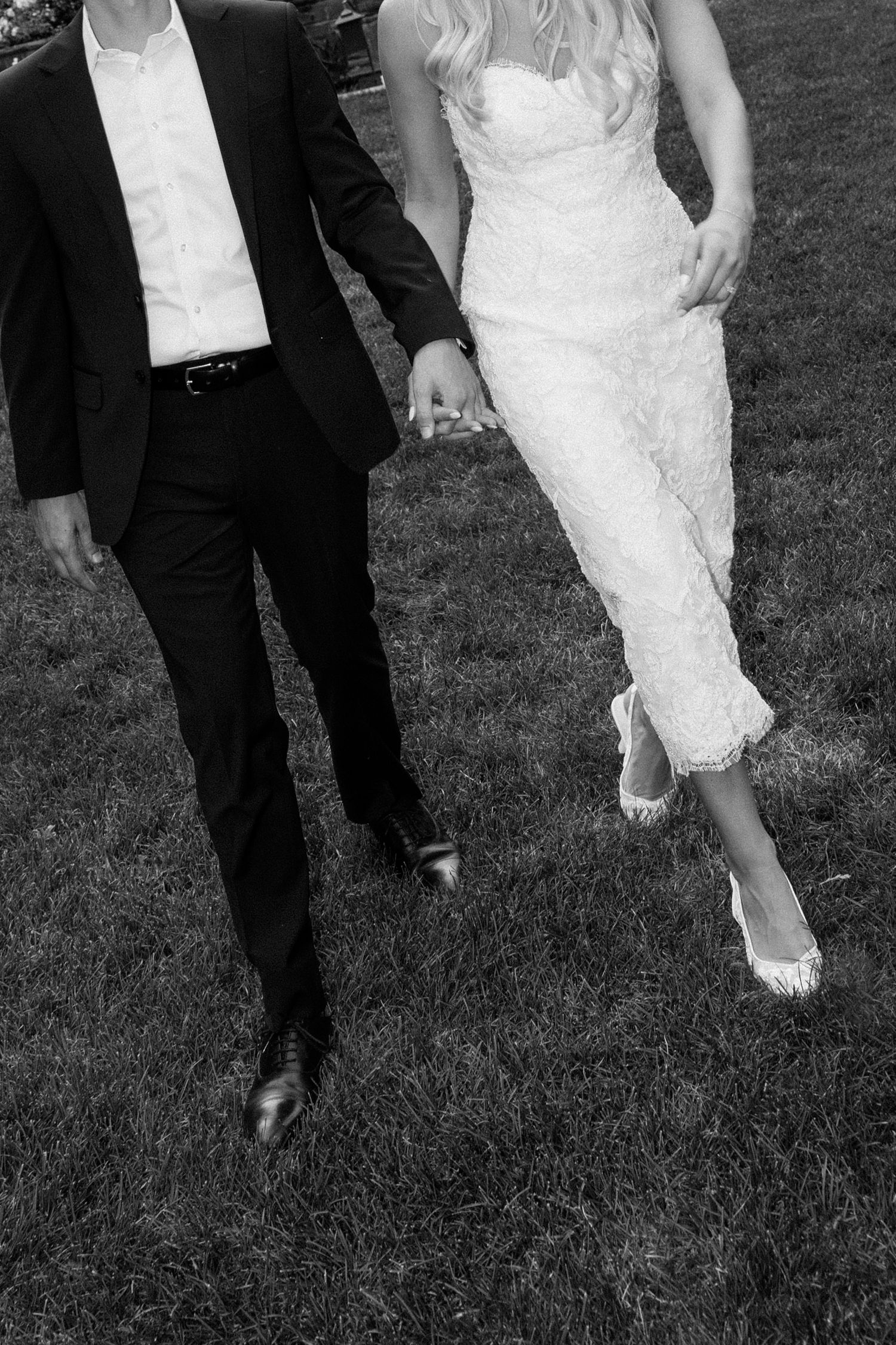 Black and white photo of bride and groom holding hands walking across green grass at wedding.