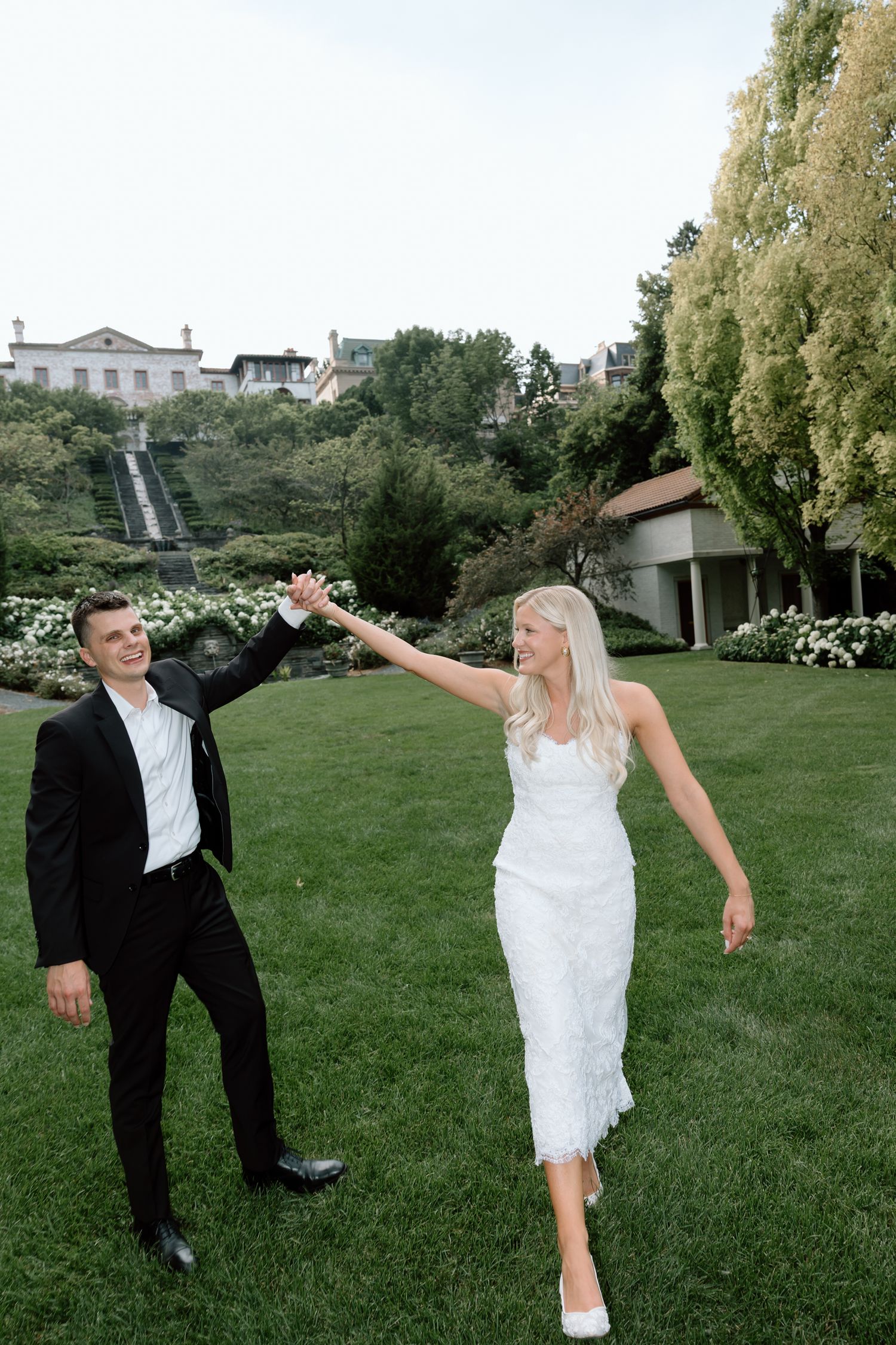 Bride and groom holding hands and dancing joyfully on green lawn with garden and estate in background.