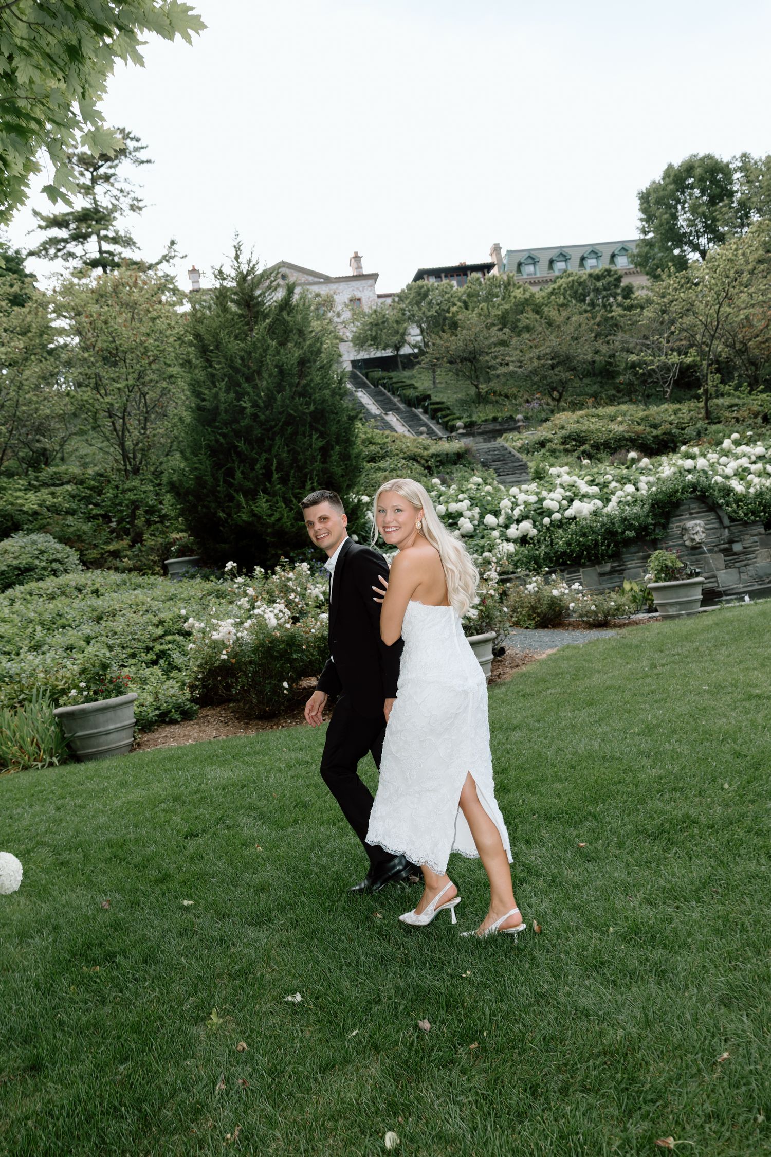 Bride and groom walking through lush garden grounds, bride glancing back in white lace midi dress.
