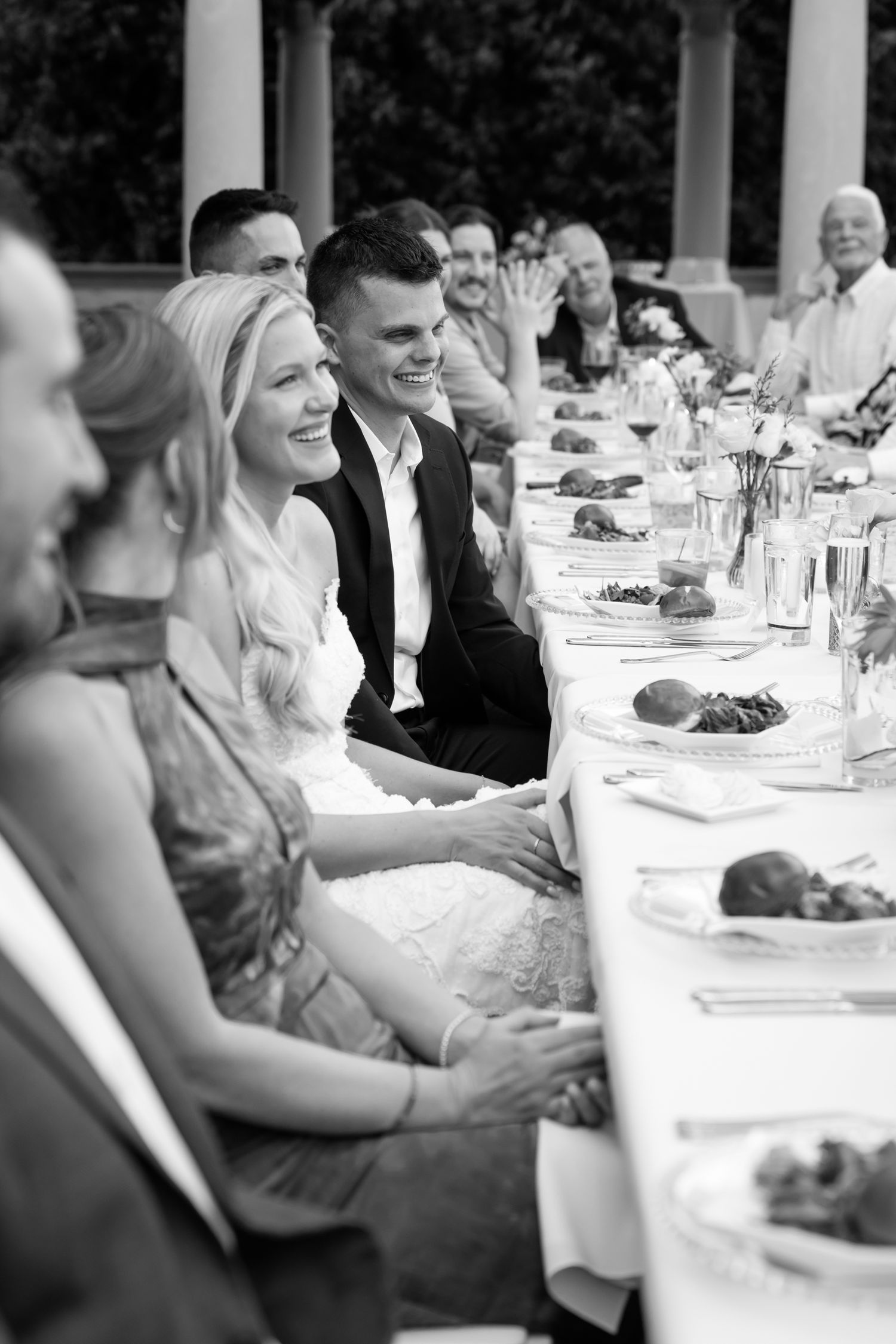 Bride and groom smiling at long outdoor wedding reception dinner table surrounded by guests.