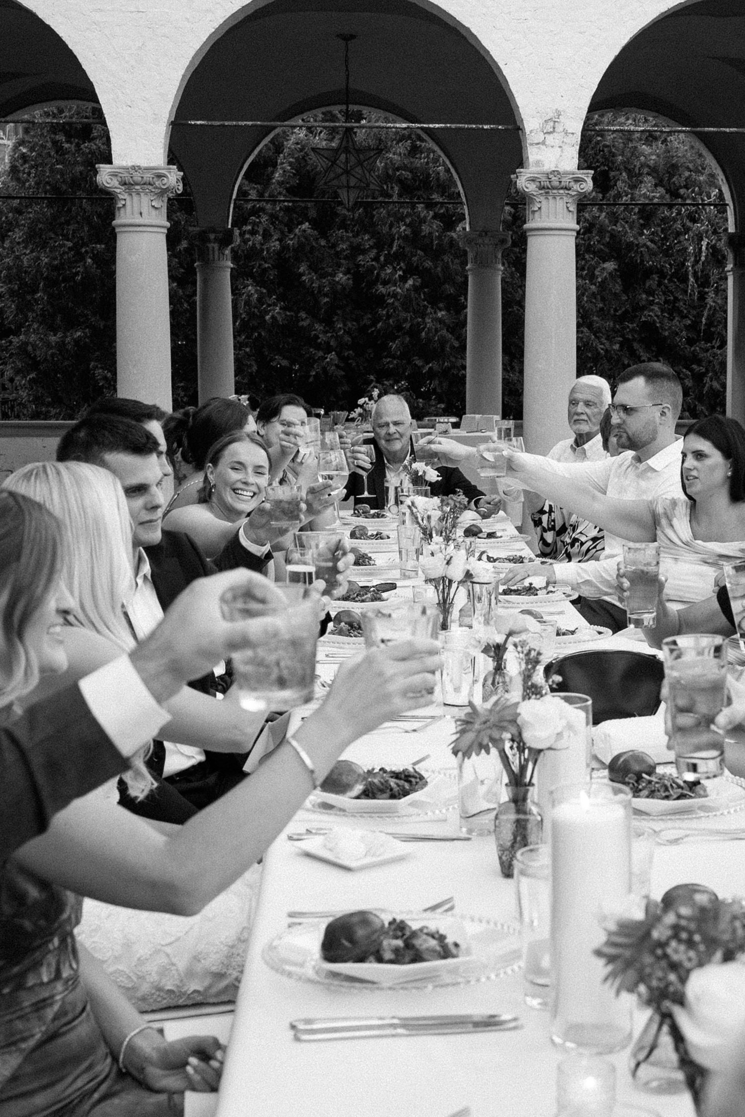 Black and white photo of wedding guests raising glasses in a toast at an elegant outdoor reception dinner under arched columns.