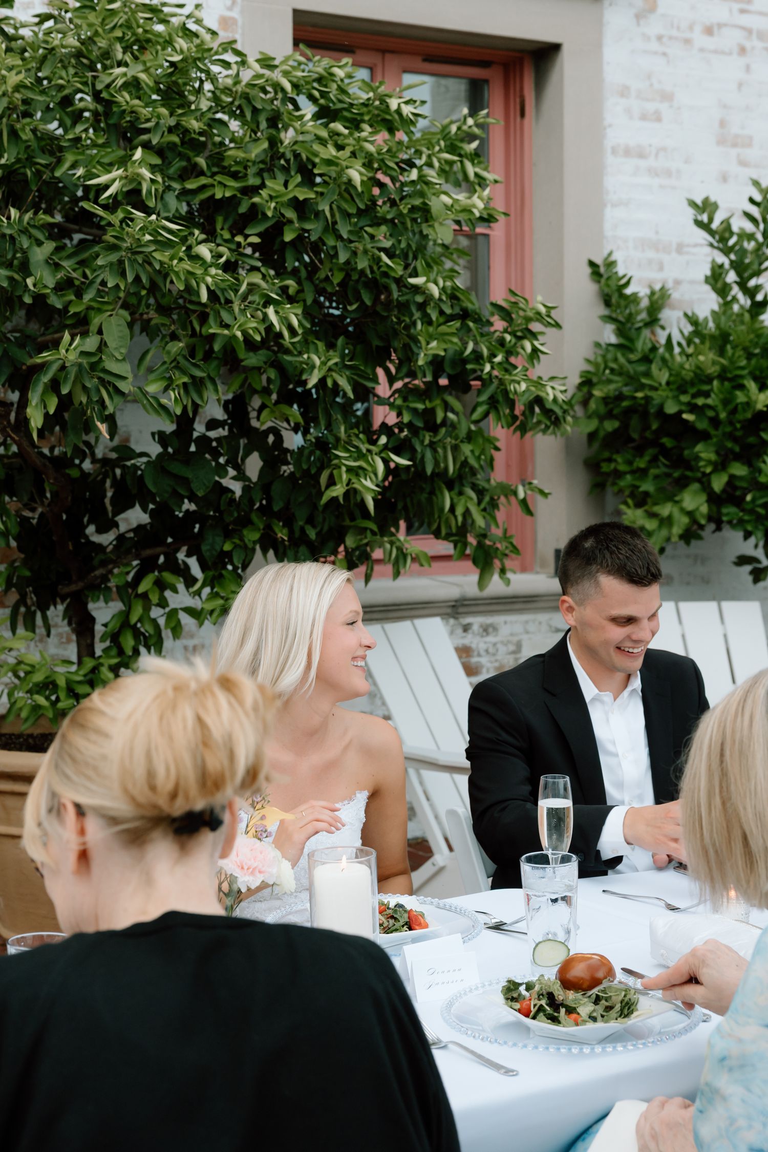 Bride and groom smiling at outdoor wedding reception dinner table with guests and champagne glasses.